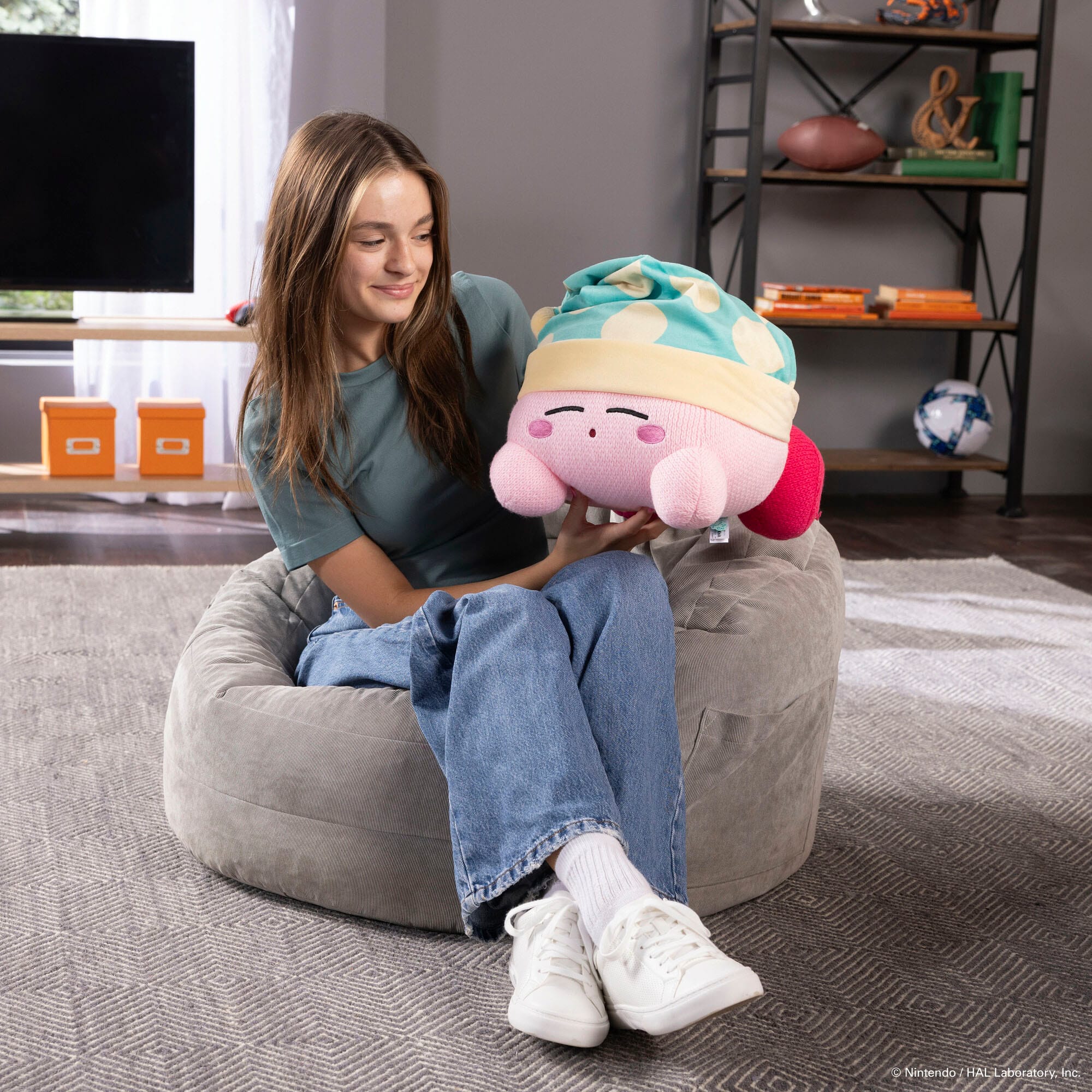 The woman is sitting on a bean bag chair and holding a stuffed animal, specifically a pink and yellow stuffed animal. She is smiling and appears to be enjoying her time with the stuffed toy. The scene takes place in a living room, with a TV in the background.
