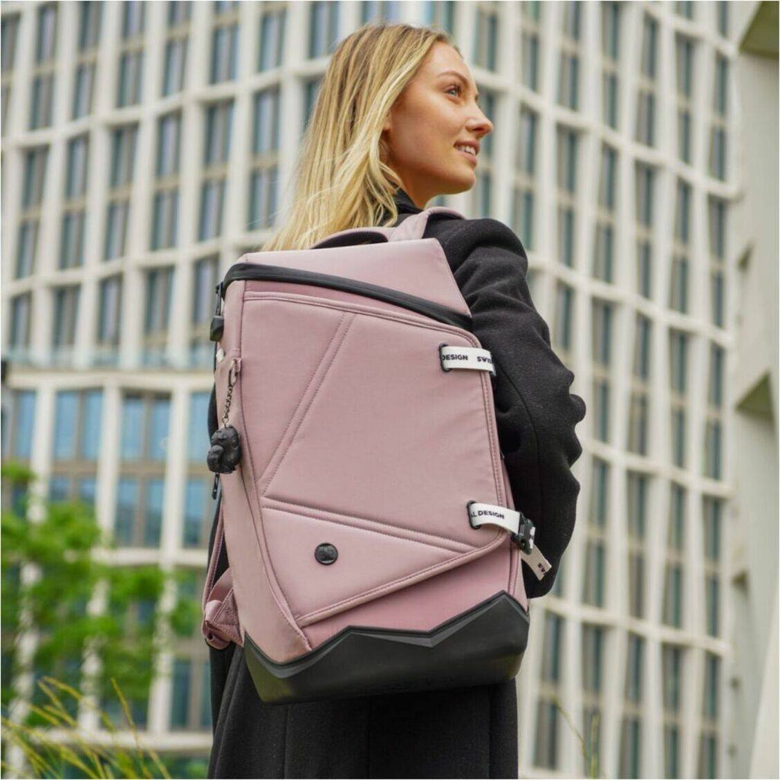 The image features a woman wearing a black jacket and carrying a pink backpack. She is standing in front of a building, possibly a skyscraper. The backpack is a prominent part of the scene, and the woman appears to be the main subject of the image.