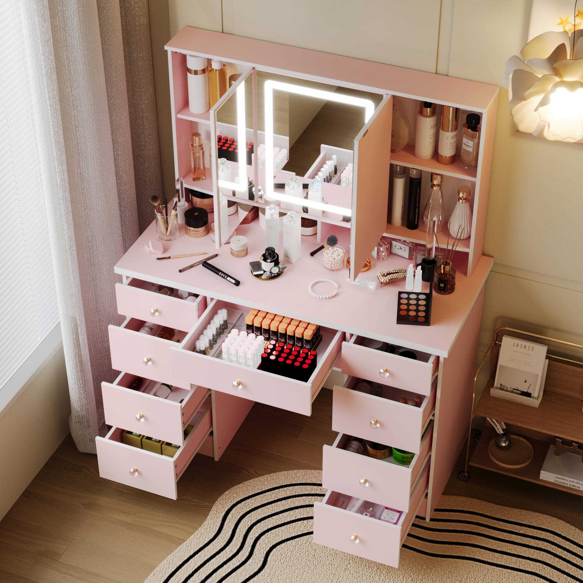 The image shows a pink vanity table with a mirror and various makeup items neatly arranged on it. The vanity has multiple drawers and shelves, with cosmetics such as lipsticks, brushes, and other beauty products organized inside and on top. The setting appears to be a well-lit, cozy room with a floral light fixture.