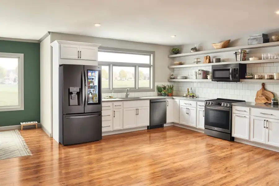 The image shows a large kitchen with wooden floors and white cabinets. The kitchen is equipped with a black refrigerator, a microwave, and an oven. There are also several bottles and bowls placed around the kitchen. The kitchen has a window, which allows natural light to enter the space. The overall appearance of the kitchen is clean and well-organized.