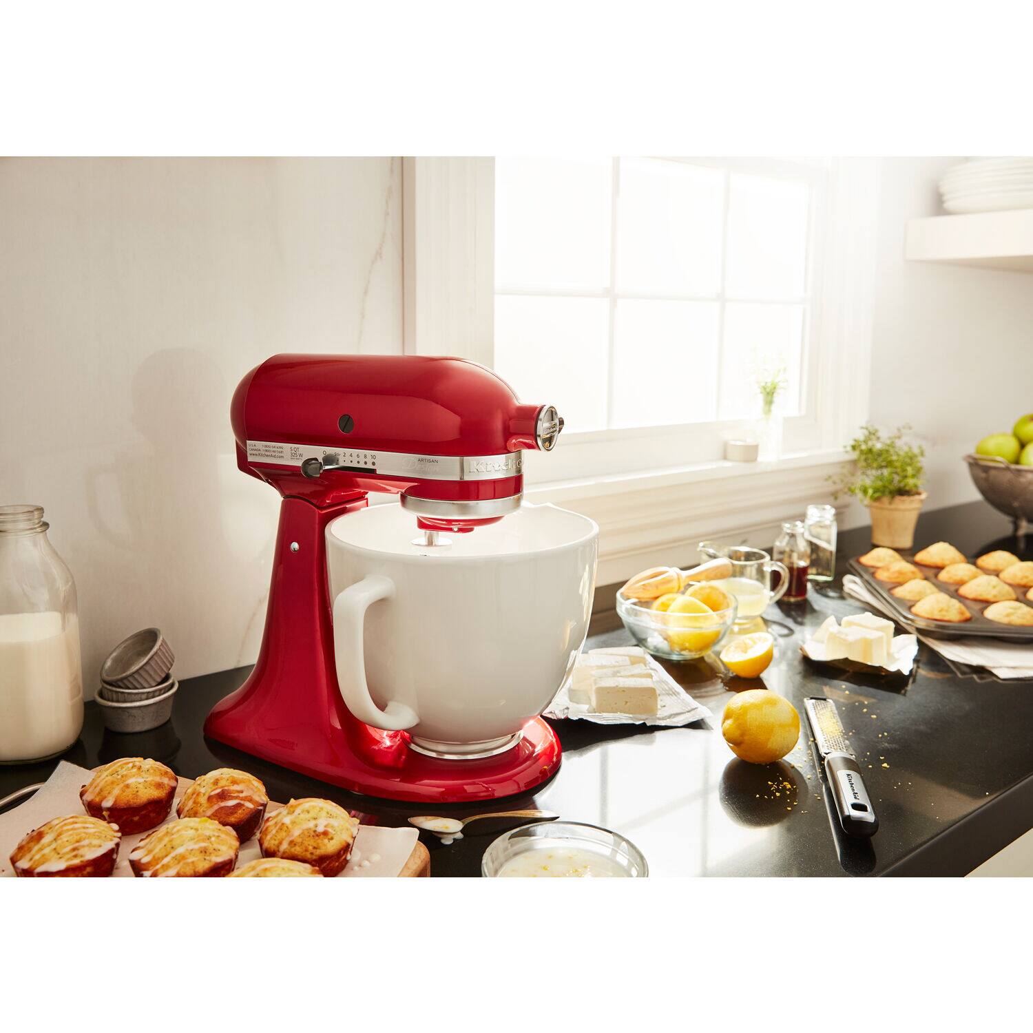 The image features a red mixer on a counter, surrounded by various food items. There are multiple cakes, a bowl, and a pile of muffins. Additionally, there are several cups and a knife on the counter. The mixer is positioned near a window, which allows natural light to illuminate the scene. The overall setting suggests a cozy and inviting atmosphere, likely in a kitchen or bakery.