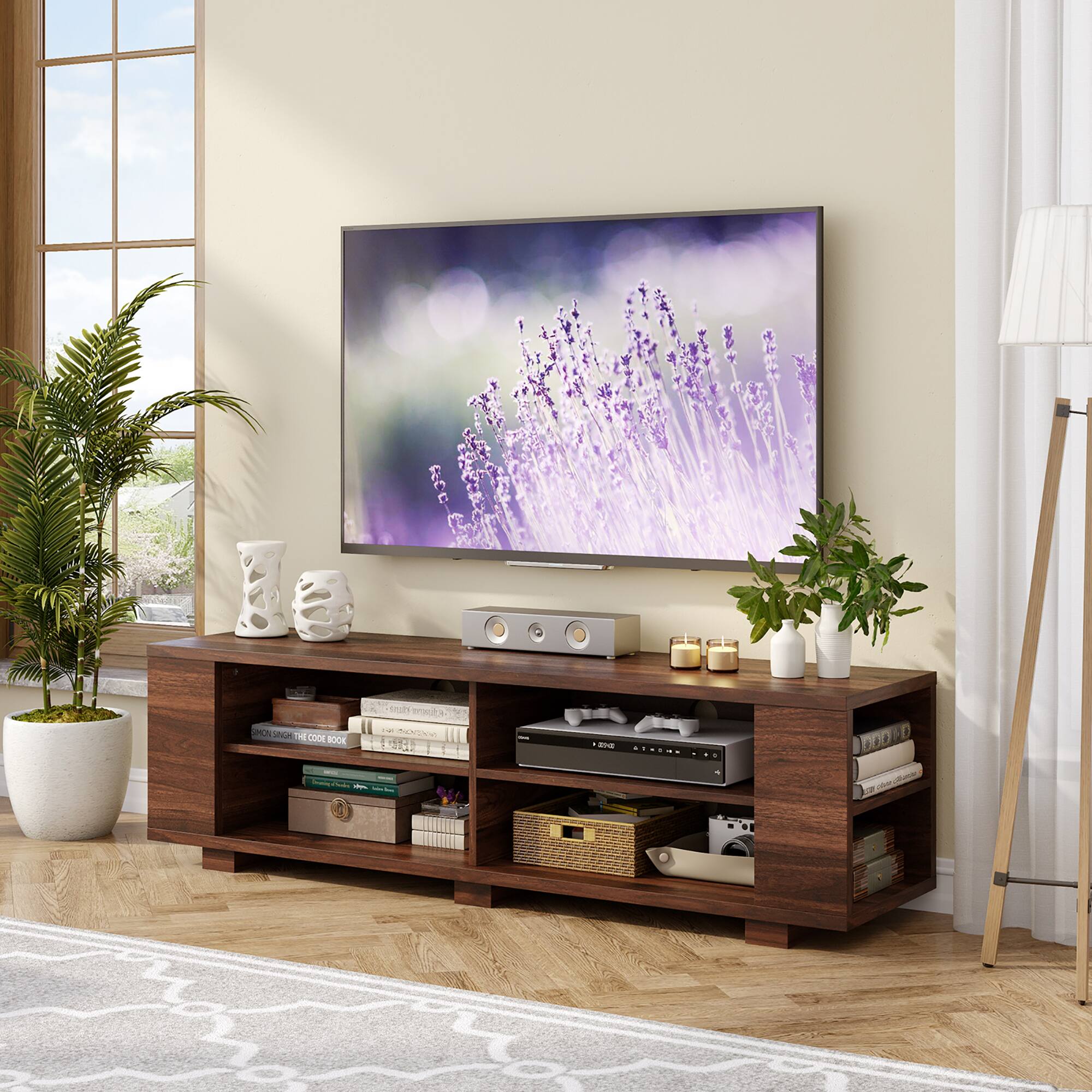 The image features a flat screen TV placed on a wooden stand in a living room. The TV is turned on, displaying a purple flower image. The stand is filled with various items, including a potted plant, a vase, and multiple books. There are also two remote controls and a keyboard on the stand. The room has a cozy and inviting atmosphere, perfect for relaxation and entertainment.