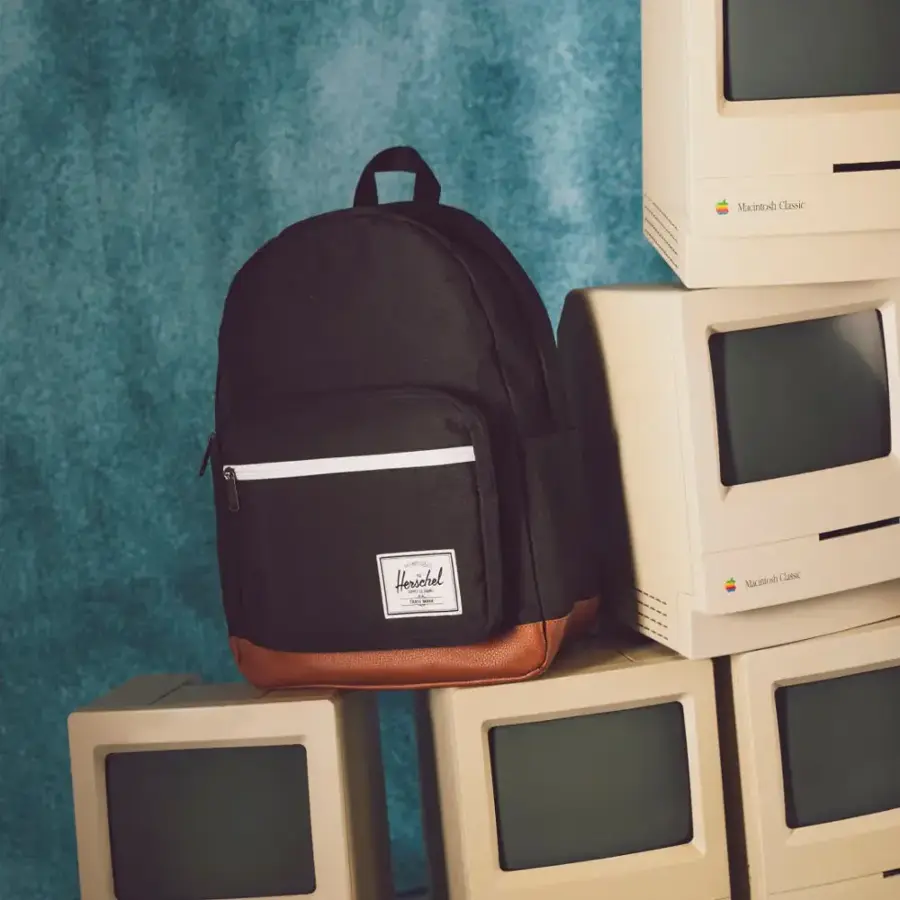 The image shows a black backpack sitting on top of a stack of old Macintosh Classic computers. The backpack is placed on the top of the stack, and the computers are arranged in a pile below it. The scene suggests a nostalgic or vintage atmosphere, as the Macintosh Classic computers are no longer in use and have been replaced by modern technology.