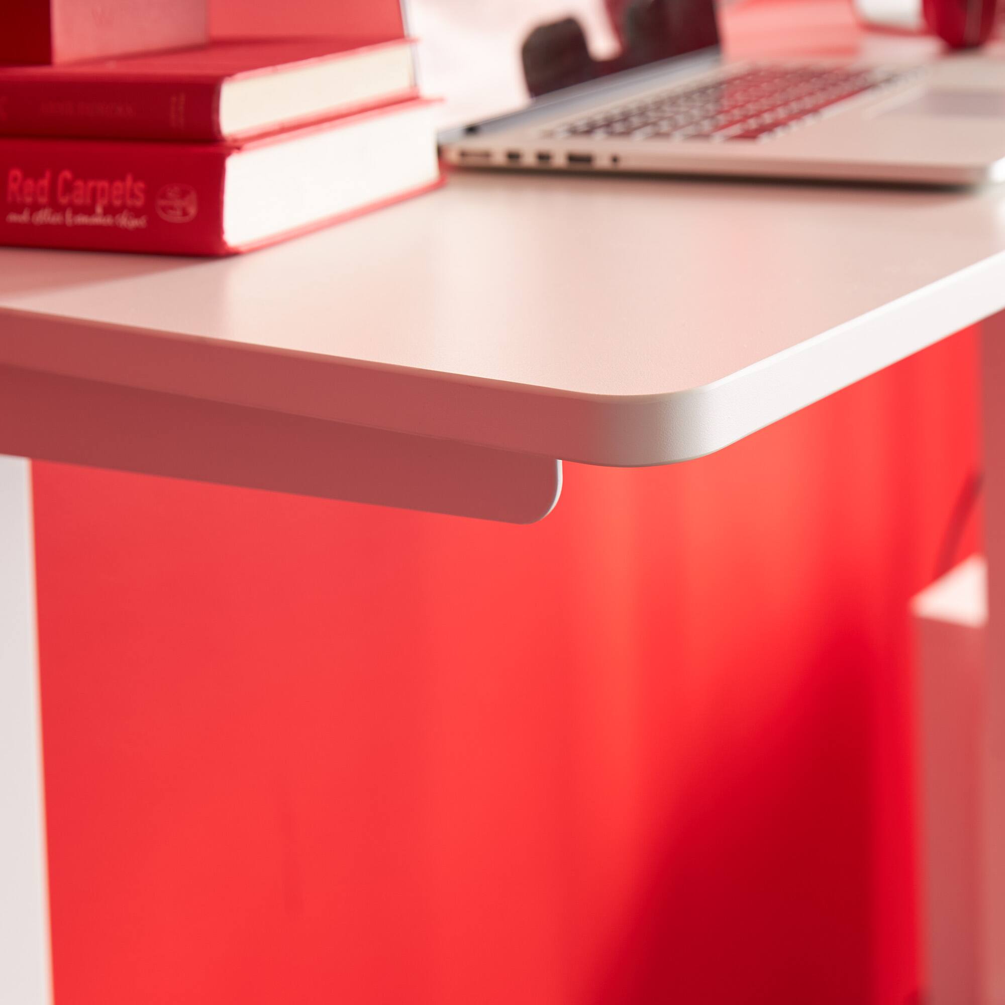 The image features a white desk with a laptop computer, a keyboard, and a mouse on it. The desk also has a few books placed on it, including a red book. The scene appears to be a workspace, possibly in a home office setting.