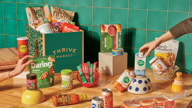 The image features a table filled with various food items, including a box of Thrive Market products. There are several bottles, a bowl, and a cup placed on the table. A person is reaching for a box of Thrive Market products, which are likely organic and healthy food options. The table is surrounded by green tiles, adding a vibrant touch to the scene.