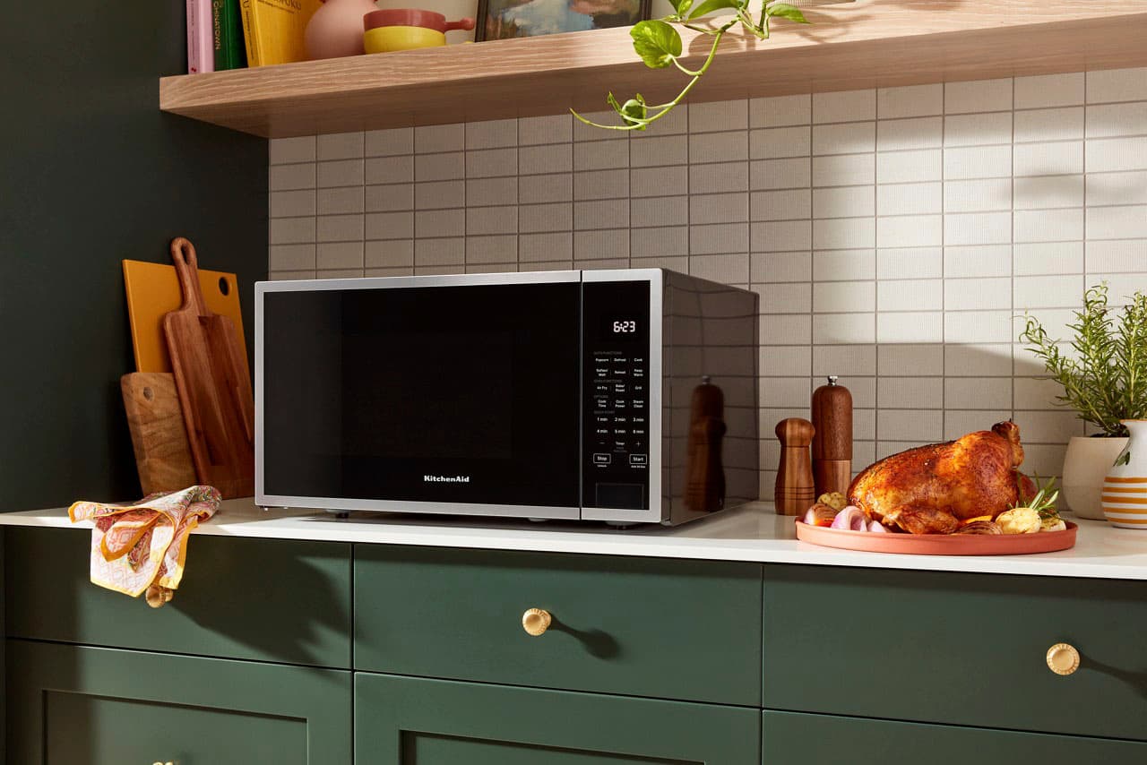 The image shows a kitchen with a microwave sitting on top of a counter. The microwave is placed in front of a tiled wall, and there are several knives and a bowl nearby. Additionally, there are two potted plants in the kitchen, one on the left side and the other on the right side. The scene appears to be in a kitchen, possibly in a home or a restaurant.