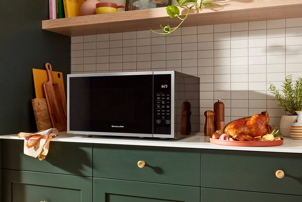 The image shows a kitchen with a microwave sitting on top of a counter. The microwave is placed in front of a tiled wall, and there are several knives and a bowl nearby. Additionally, there are two potted plants in the kitchen, one on the left side and the other on the right side. The scene appears to be in a kitchen, possibly in a home or a restaurant.