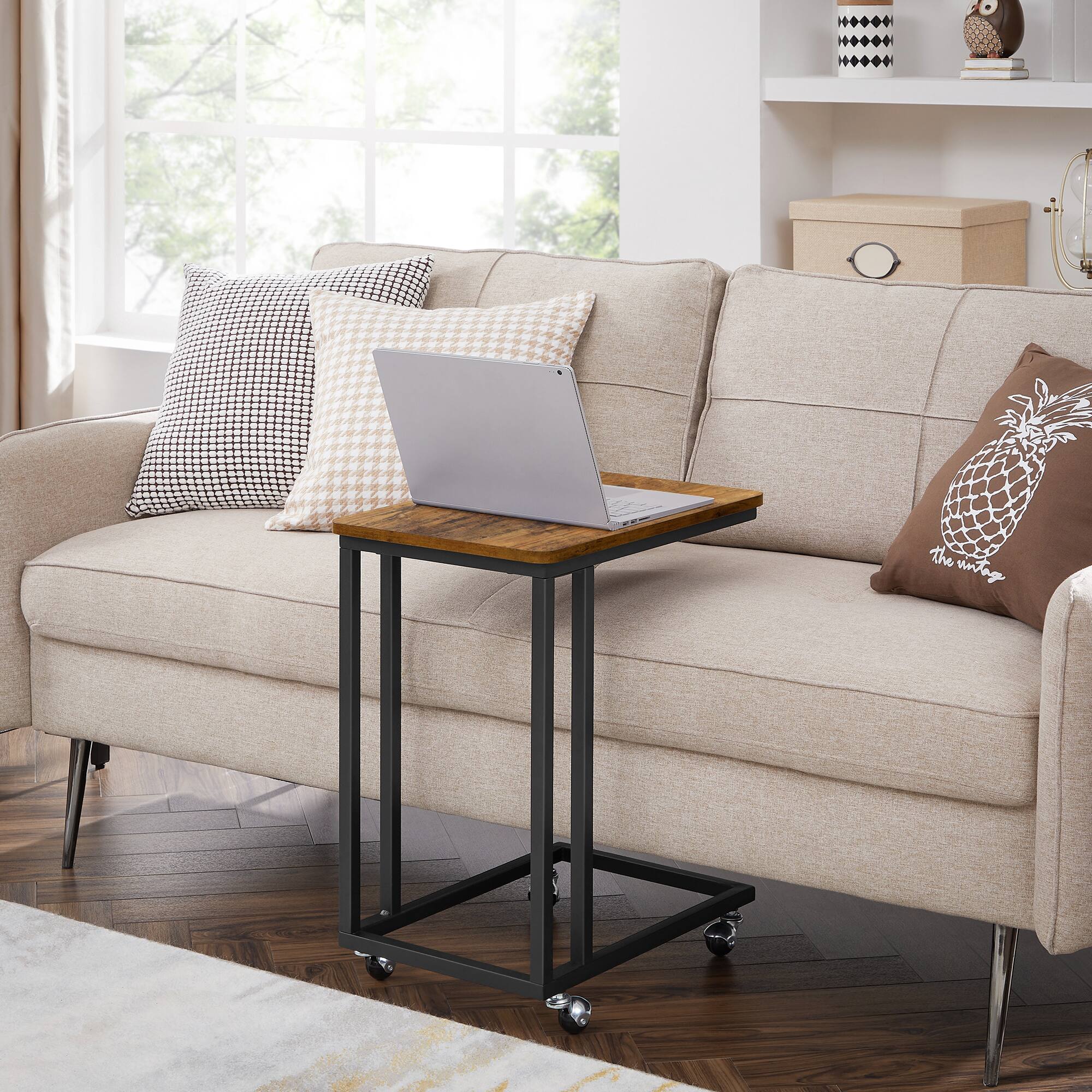 The image shows a cozy living room setting with a beige sofa. On the sofa, there are two patterned pillows: one with a black and white geometric design and another with a beige and white geometric pattern. A laptop is placed on a black metal side table with a wooden top. To the right of the sofa, there is a brown pillow with a pineapple design and the text "The Unravel." In the background, there is a wooden chest and a shelf with decorative items. The room has large windows with white curtains, allowing natural light to fill the space.