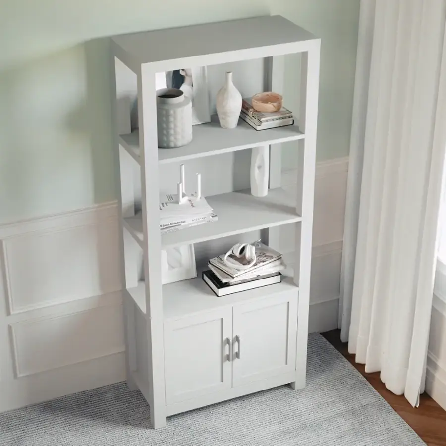 The image features a white bookshelf with various items on it. There are several books placed on different shelves, as well as a vase and a bowl. The bookshelf is situated in a room with a window nearby, and it is placed on a rug. The arrangement of the items on the shelves creates a visually appealing and organized display.