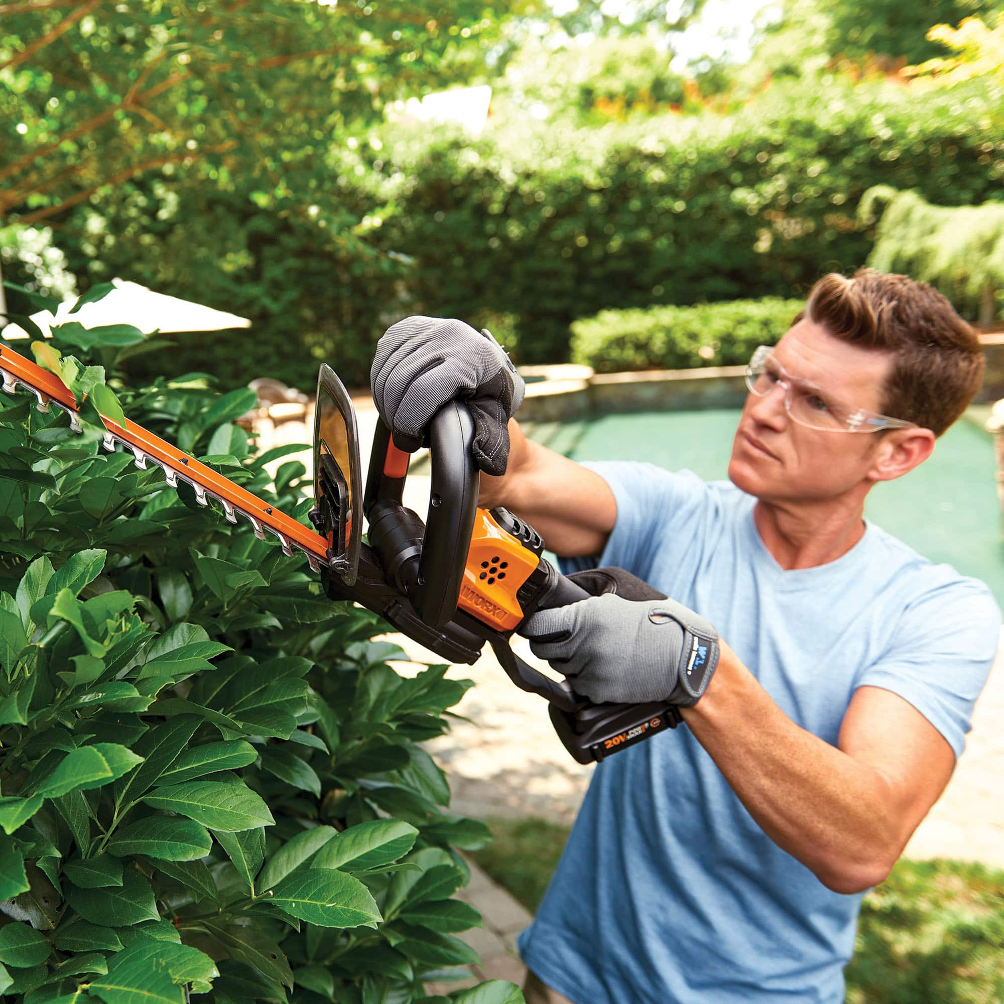 The man in the image is wearing a blue shirt and is using a large pair of scissors to trim a bush. He is standing in a backyard, surrounded by greenery. The scissors are quite large, and the man is focused on his task.