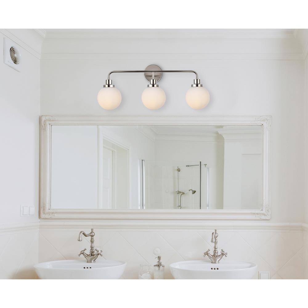 The image shows a bathroom with a large mirror above two white sinks. The mirror has a decorative frame, and there are three round light fixtures above it. The sinks have ornate silver faucets, and the walls are painted white.