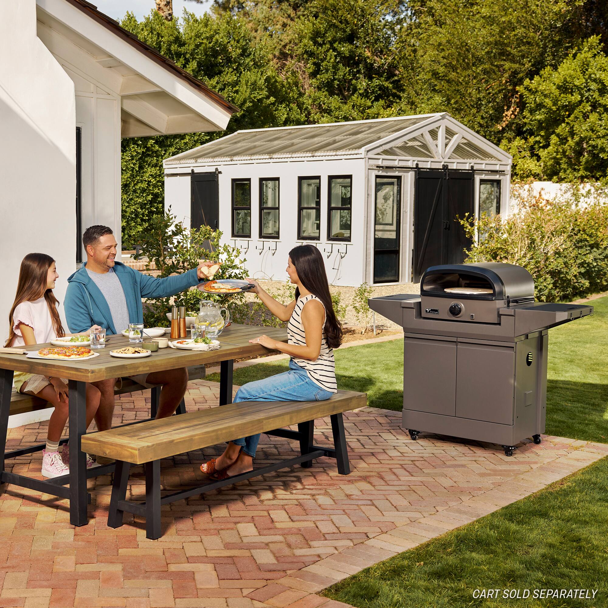 The image shows a family gathered around a wooden dining table, enjoying a meal together. There are four people in the scene, with two adults and two children. The table is set with various items such as cups, a bowl, and a pizza. A bench is placed near the table, providing additional seating.

In the background, there is a grill, indicating that the family might be having a barbecue or outdoor meal. The scene captures a moment of togetherness and bonding between the family members.