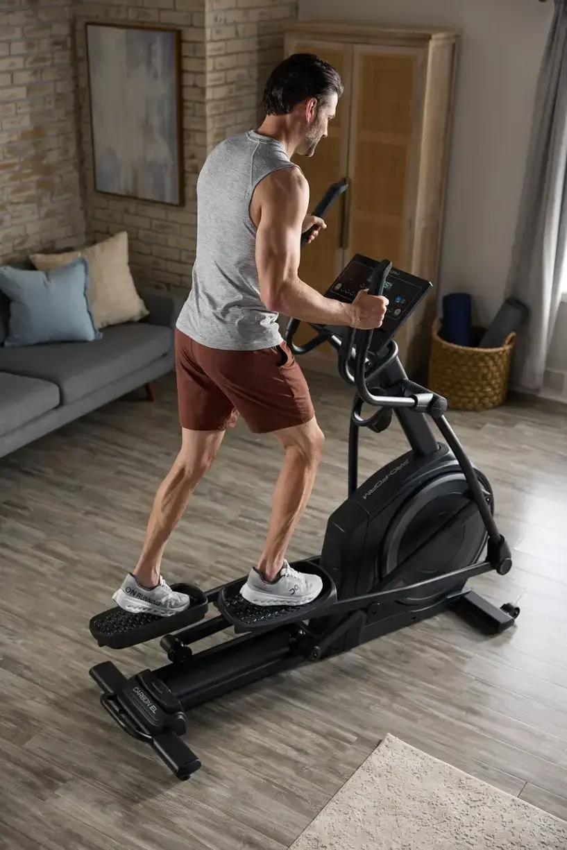The image shows a man using a Pro-Form Mad On Runner DC Carbon El treadmill in his living room. He is wearing shorts and is actively running on the treadmill. The living room has a couch and a chair, and there is a TV mounted on the wall. The man is focused on his workout, and the treadmill provides a convenient way to exercise indoors.