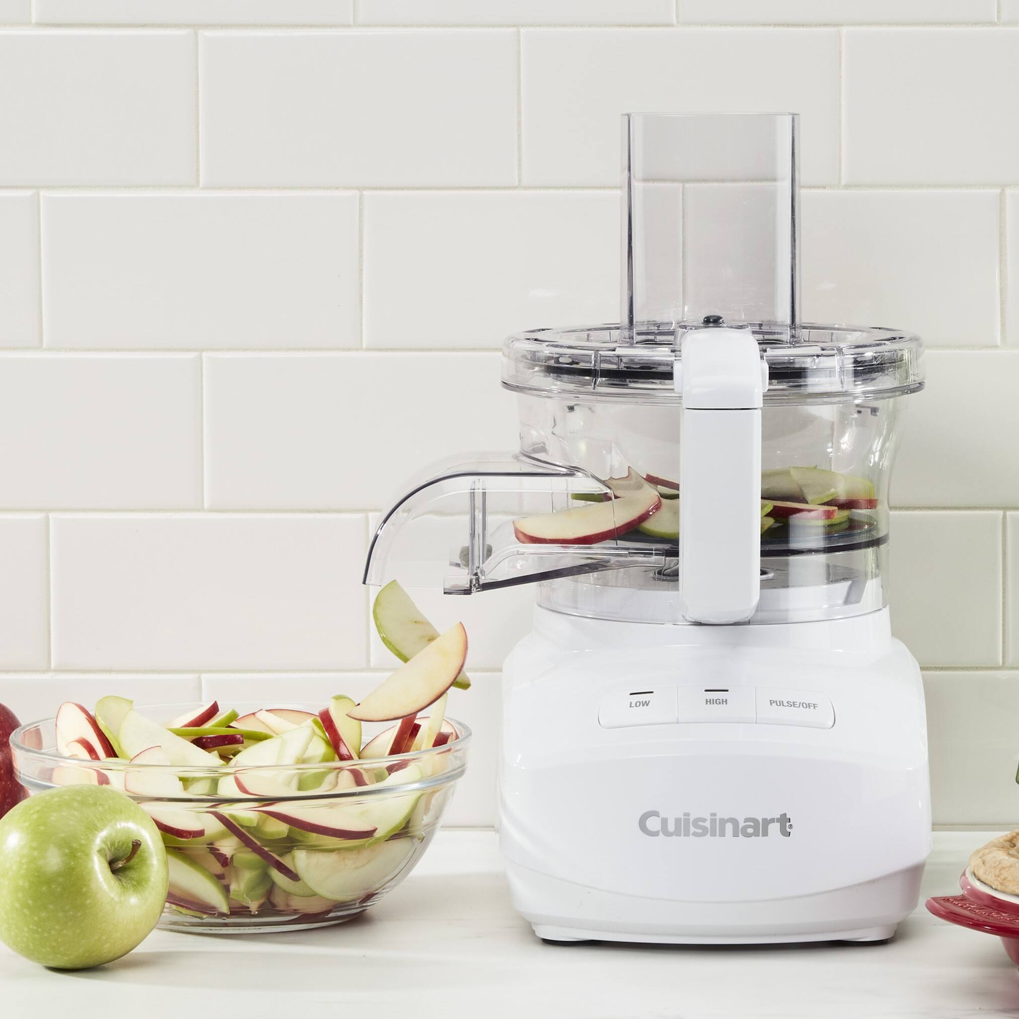 The image features a Cuisinart food processor on a counter, surrounded by various fruits and vegetables. There are apples, oranges, and broccoli nearby, as well as a bowl of apples and a bowl of oranges. The food processor is placed next to a tiled wall, and the scene is set against a white background.