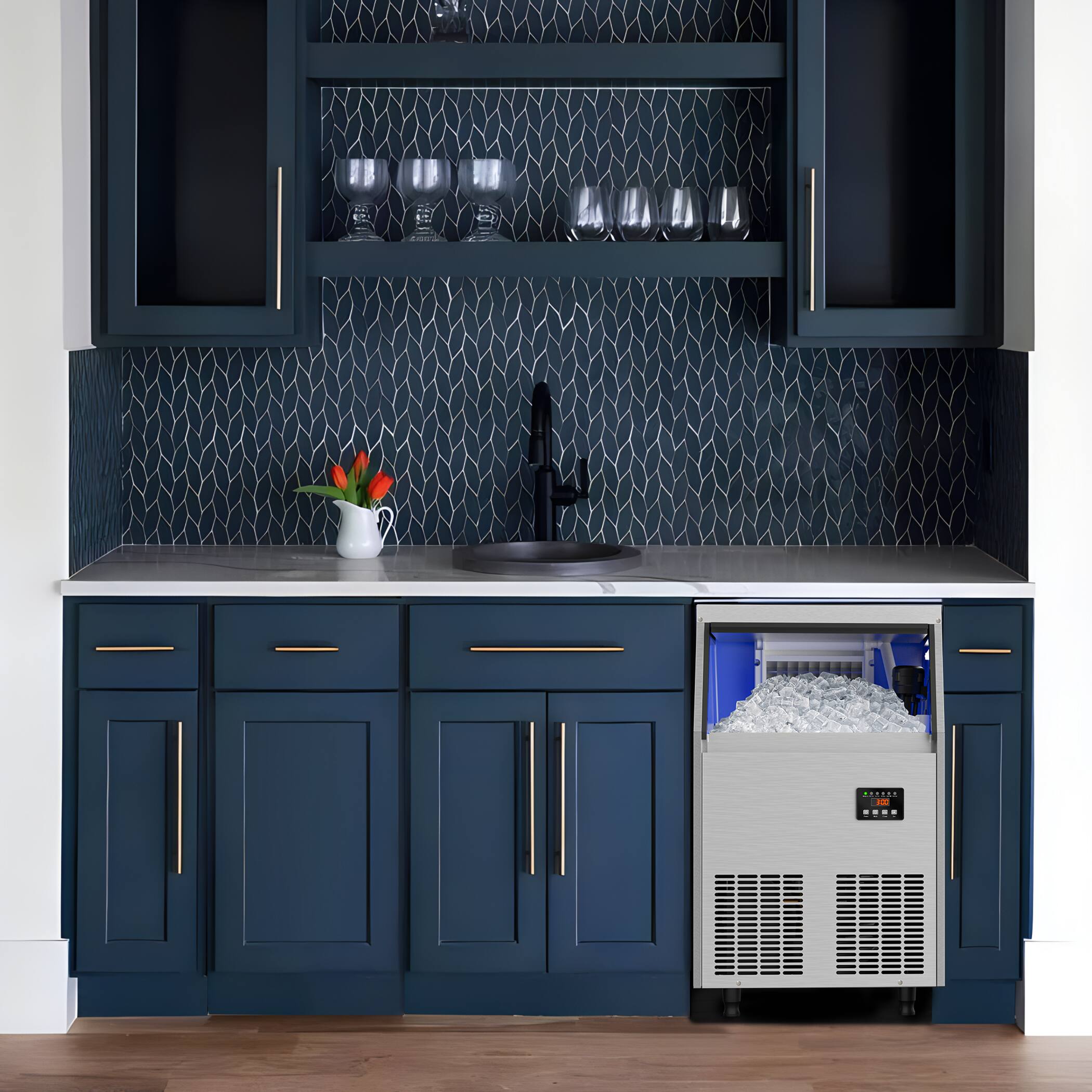 The image shows a modern kitchen with dark blue cabinets and gold handles. The backsplash features a geometric pattern. On the countertop, there is a white vase with orange flowers and a black faucet. Below the countertop, there is a built-in ice maker with a digital display. The kitchen has a sleek and contemporary design.