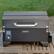 Cuisinart is the brand of the grill shown in the image. It is a black grill with a silver handle and is sitting on a wooden table.