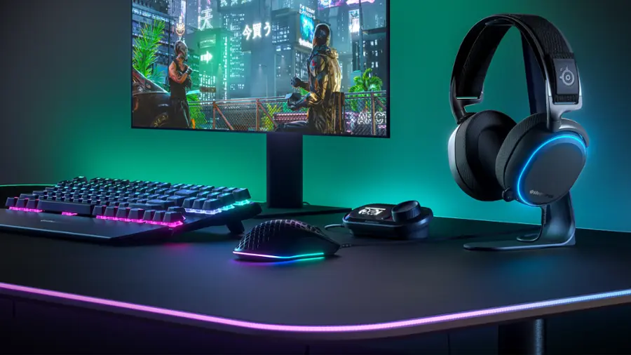 The image features a desk with a computer setup, including a keyboard, a mouse, and a monitor. The monitor is turned on, displaying a video game. There is also a pair of headphones placed on the desk, likely for an immersive gaming experience. The desk is illuminated with purple and blue lights, creating a visually appealing workspace.