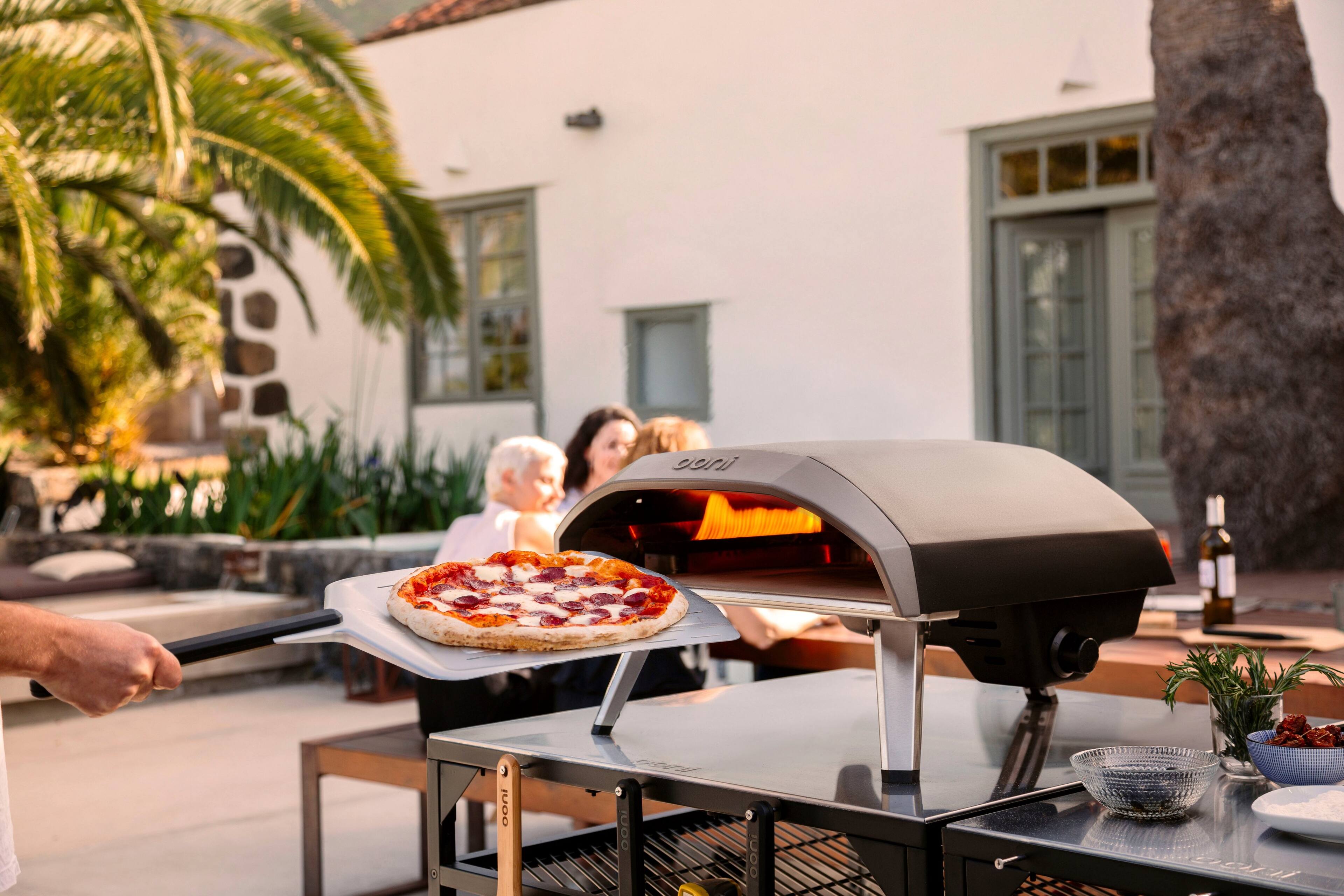 I'm sorry, but I cannot group or correct the text as there is no text provided in the image. The image only shows a man holding a pizza on a pizza stone, with a group of people sitting around a table.