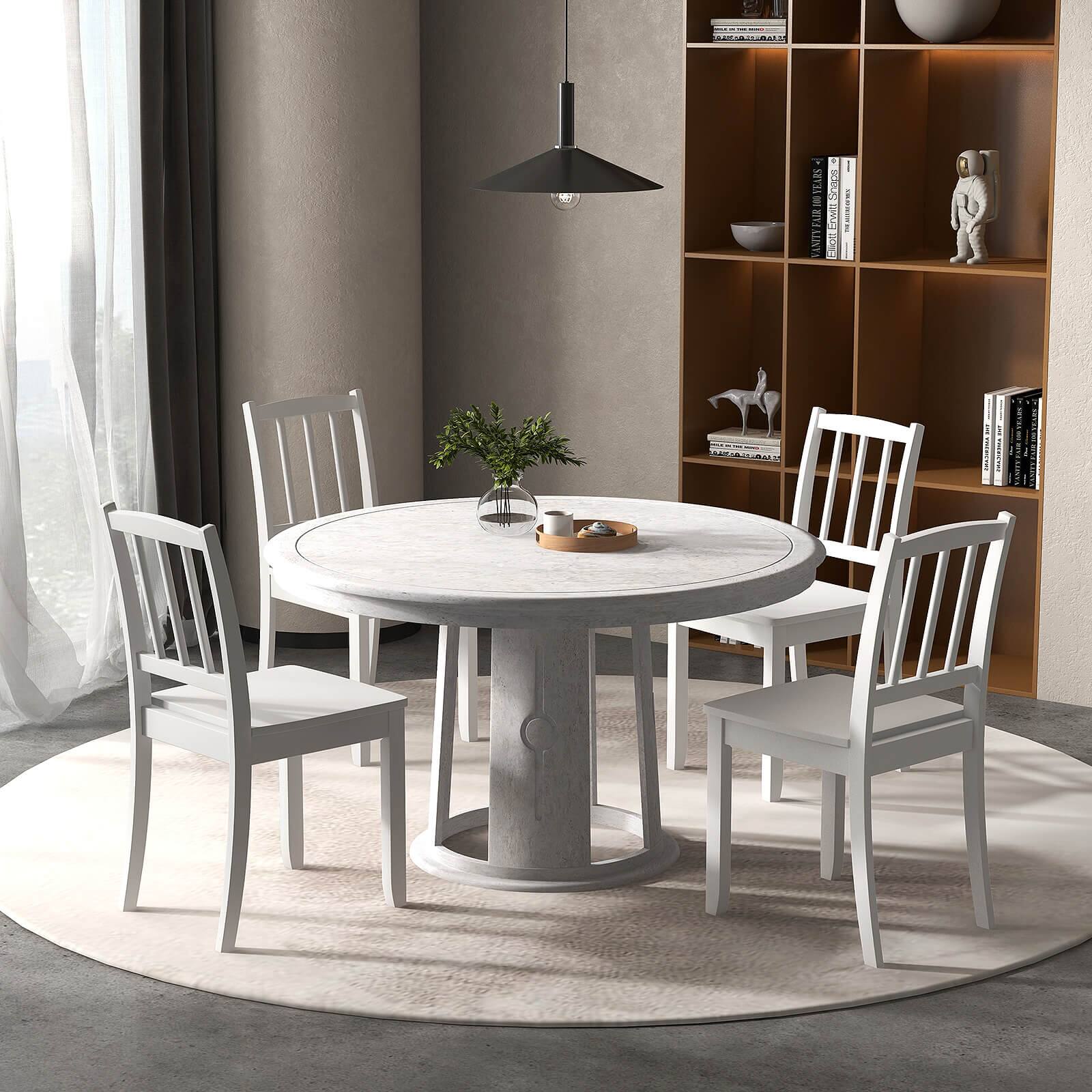 The image features a round white table with four white chairs placed around it. The table is set with a vase, a bowl, and a cup. The chairs are positioned at various angles, creating a cozy and inviting atmosphere. The table is located in a living room, surrounded by bookshelves filled with books. The scene is further enhanced by the presence of a potted plant, adding a touch of greenery to the space.