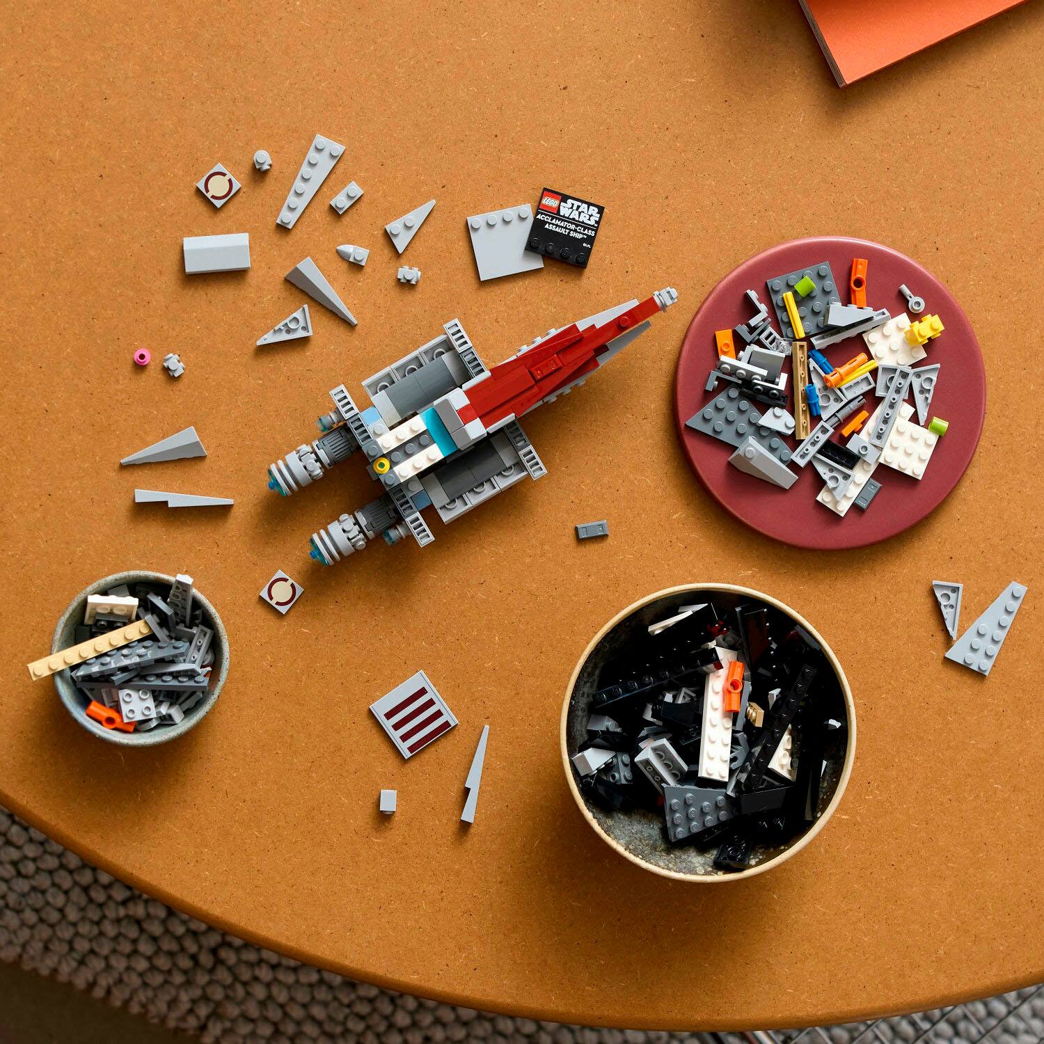 The image shows a table with a bowl of lego pieces, a cup of lego pieces, and a lego spaceship. The table is made of wood and has a red surface. The lego spaceship is the main focus of the scene, and it appears to be a large model. The table is also surrounded by other items, such as a book and a cup.
