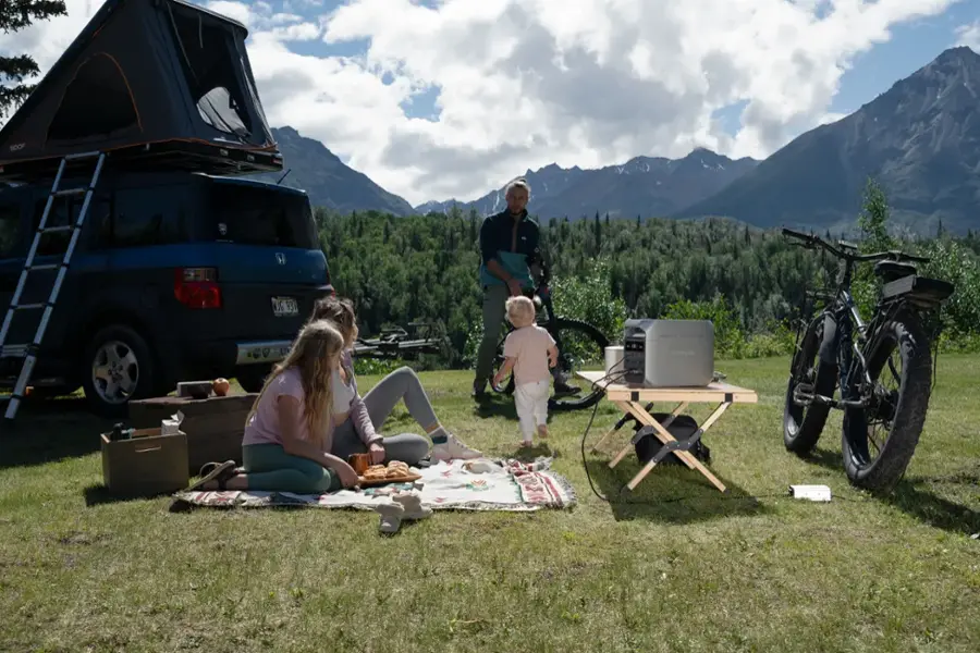 The image shows a group of people sitting on a blanket in a grassy field, with a picnic table nearby. There are two bicycles and a motorcycle parked nearby, as well as a car and a truck. The people seem to be enjoying their time outdoors, possibly on a camping trip. The presence of the bicycles and motorcycle suggests that they might be engaging in outdoor activities, such as cycling or exploring the area. The car and truck indicate that they have brought their belongings and supplies for the trip, making it a well-planned and organized outing.