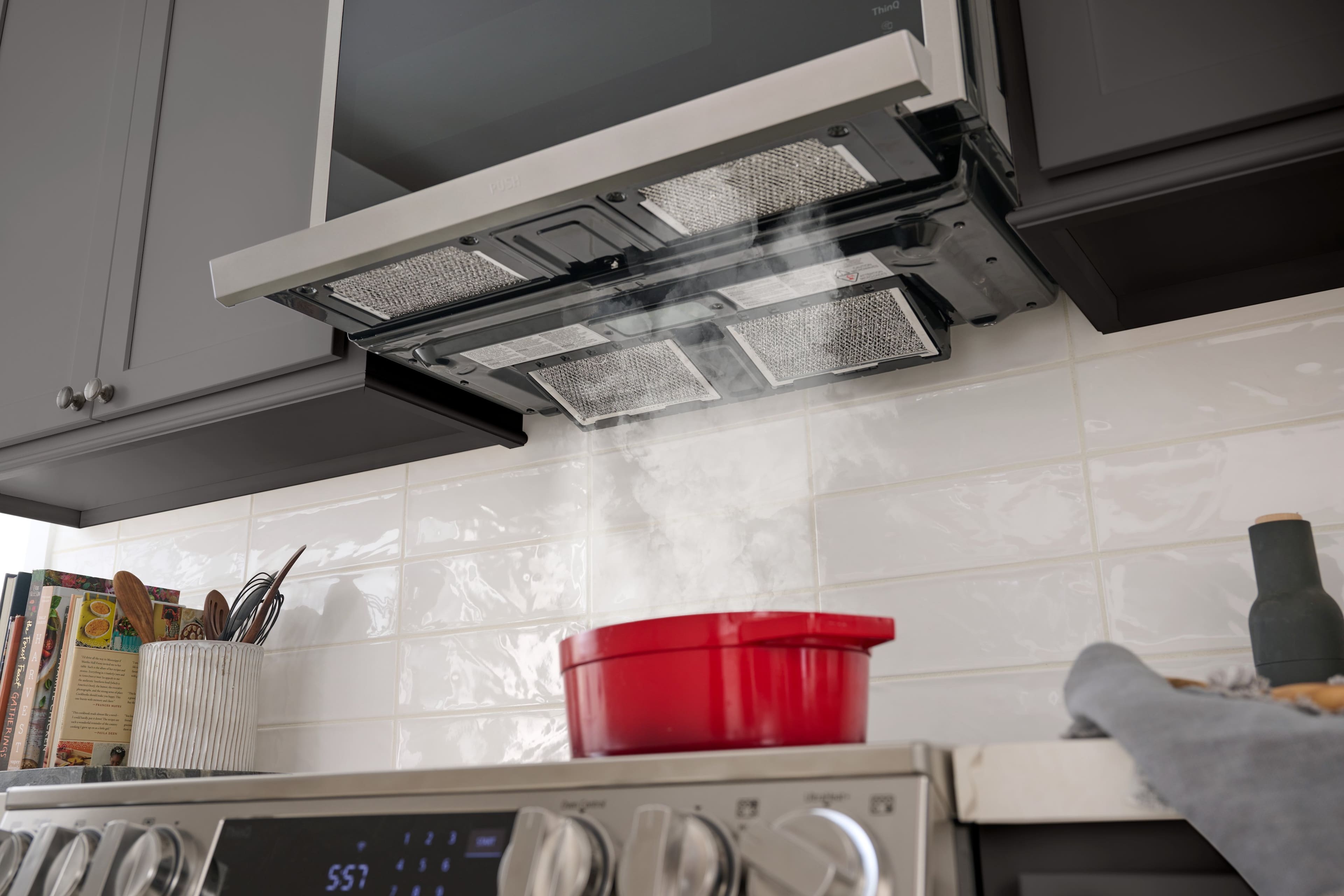 The image shows a kitchen with a stove and a microwave. The stove has a pot on it, and the microwave is located above the stove. The oven is also present in the kitchen, with a timer showing the time of 5:08. The kitchen is equipped with various utensils such as knives, spoons, and a bowl. There are also several books placed in the kitchen, possibly cookbooks or other reading materials.