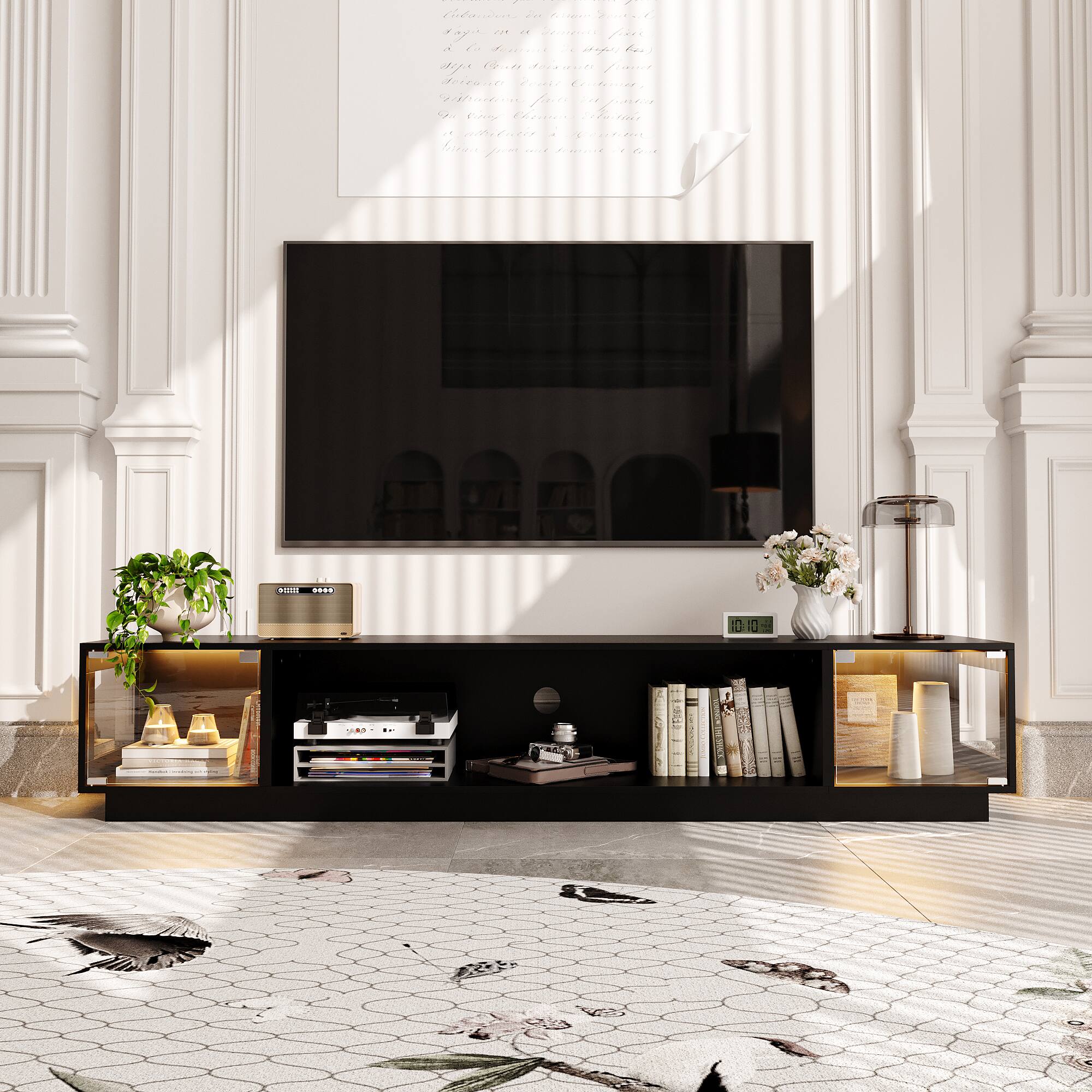 I am sitting in a room with a flat screen TV on the wall. There are two speakers on the table, and a large bookshelf filled with books. The room also has a potted plant and a vase with flowers. The sunlight is shining through the window, creating a bright and inviting atmosphere.