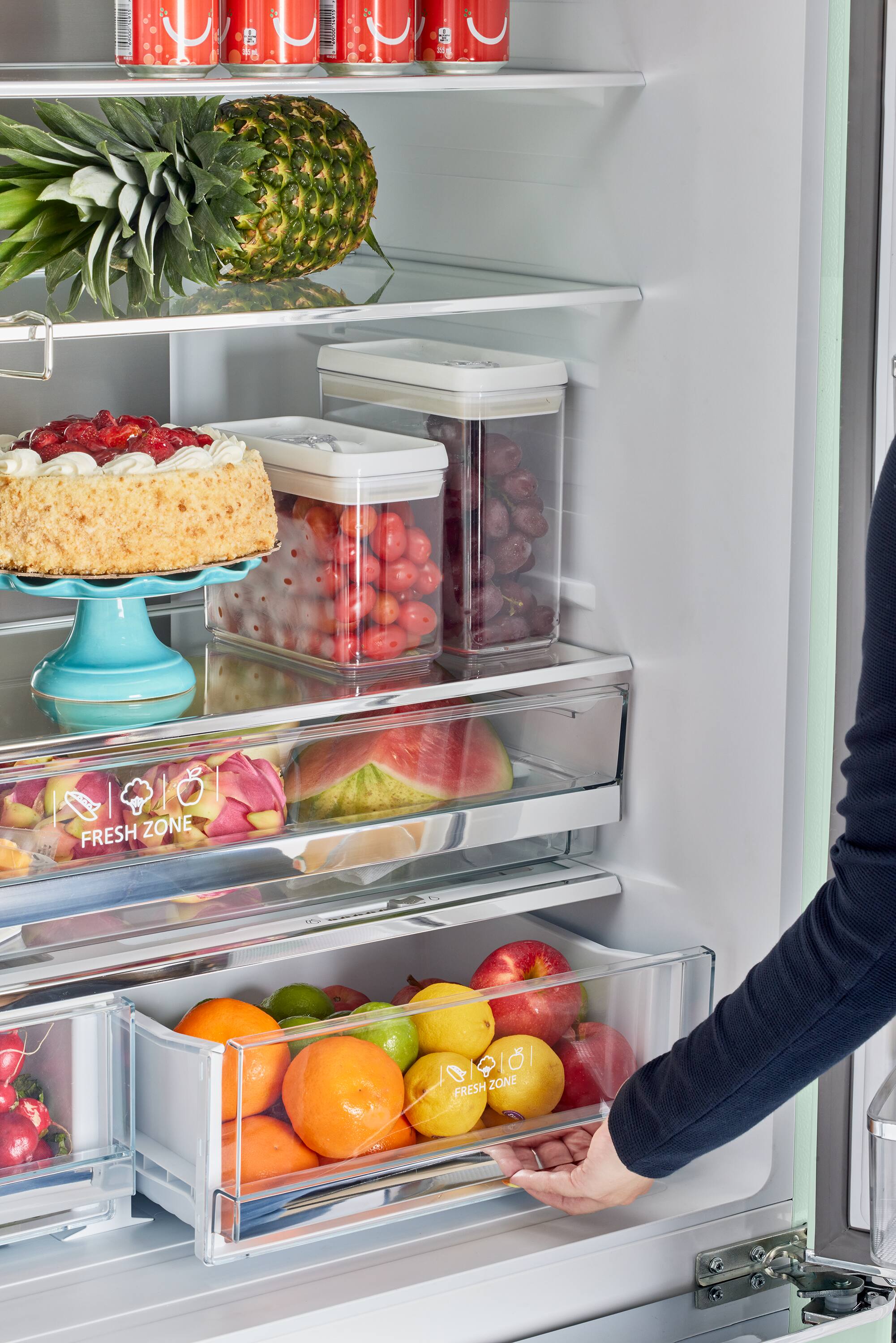 The image shows a refrigerator with a variety of food items, including fruits and vegetables. The person is reaching for a container of fruit in the refrigerator. The refrigerator has a fresh zone, which is indicated by the presence of fruits and vegetables. The image also shows a person's hand holding a container of fruit, and the refrigerator is filled with food items.