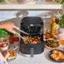 The image shows a person using a food processor to prepare a meal. The food processor is filled with various ingredients, including broccoli, carrots, and potatoes. The person is holding the food processor handle, and the machine is open, revealing the food inside.

In addition to the food processor, there are several other items on the counter, such as a bowl, a fork, a knife, a spoon, and a bottle. There are also a few oranges and a potted plant nearby, adding to the overall ambiance of the scene.