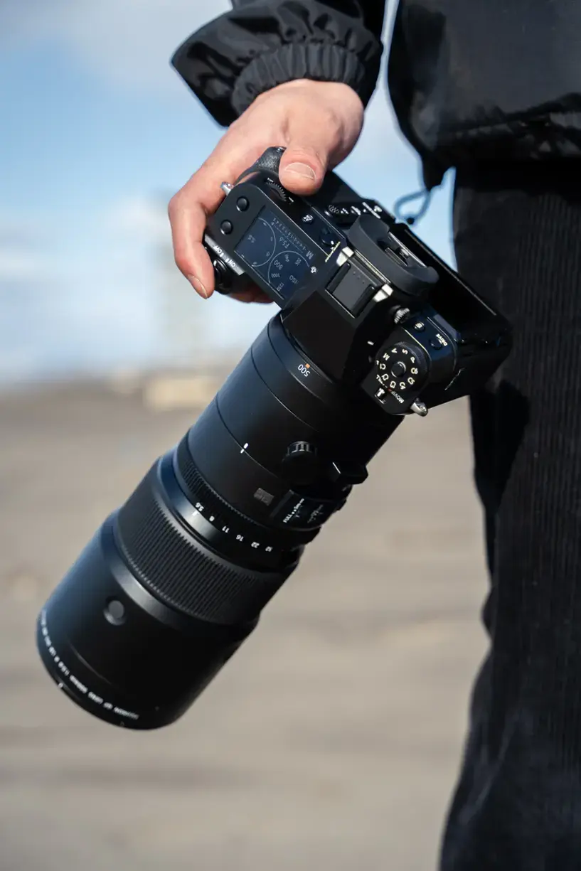 The image features a person holding a large camera lens, which is likely a telephoto lens. The lens is being held in front of the person, and it appears to be a professional-grade camera lens. The person is wearing a black jacket, which adds to the overall atmosphere of the scene.