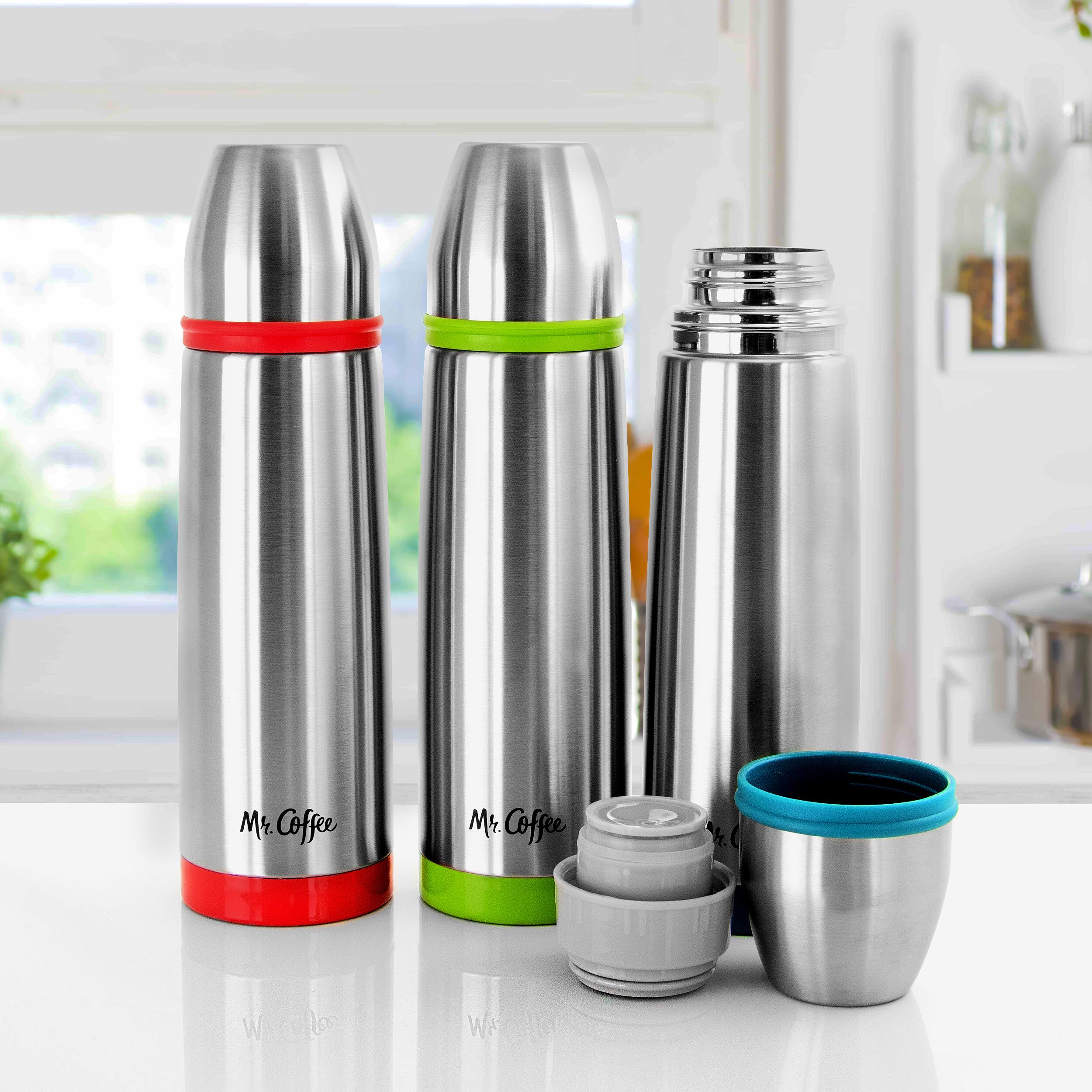 Mr. Coffee is a brand of coffee makers and coffee-related accessories. In the image, there are three Mr. Coffee coffee canisters, each with a different color: red, green, and silver. They are accompanied by a blue coffee cup and a silver coffee mug. The image also features a potted plant and a window in the background.