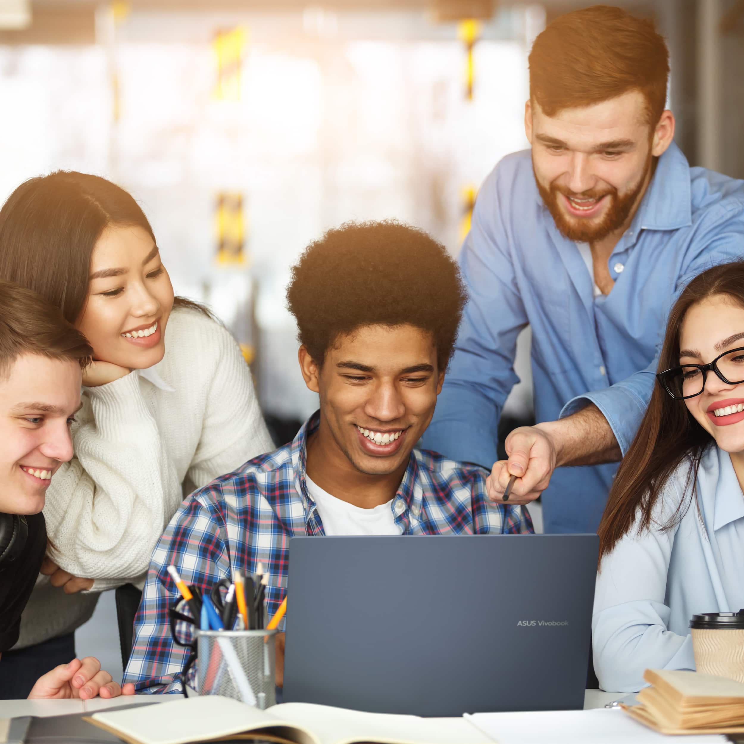 The group of people is gathered around a laptop computer, which is an ASUS Vivobook. They are all smiling and enjoying their time together.
