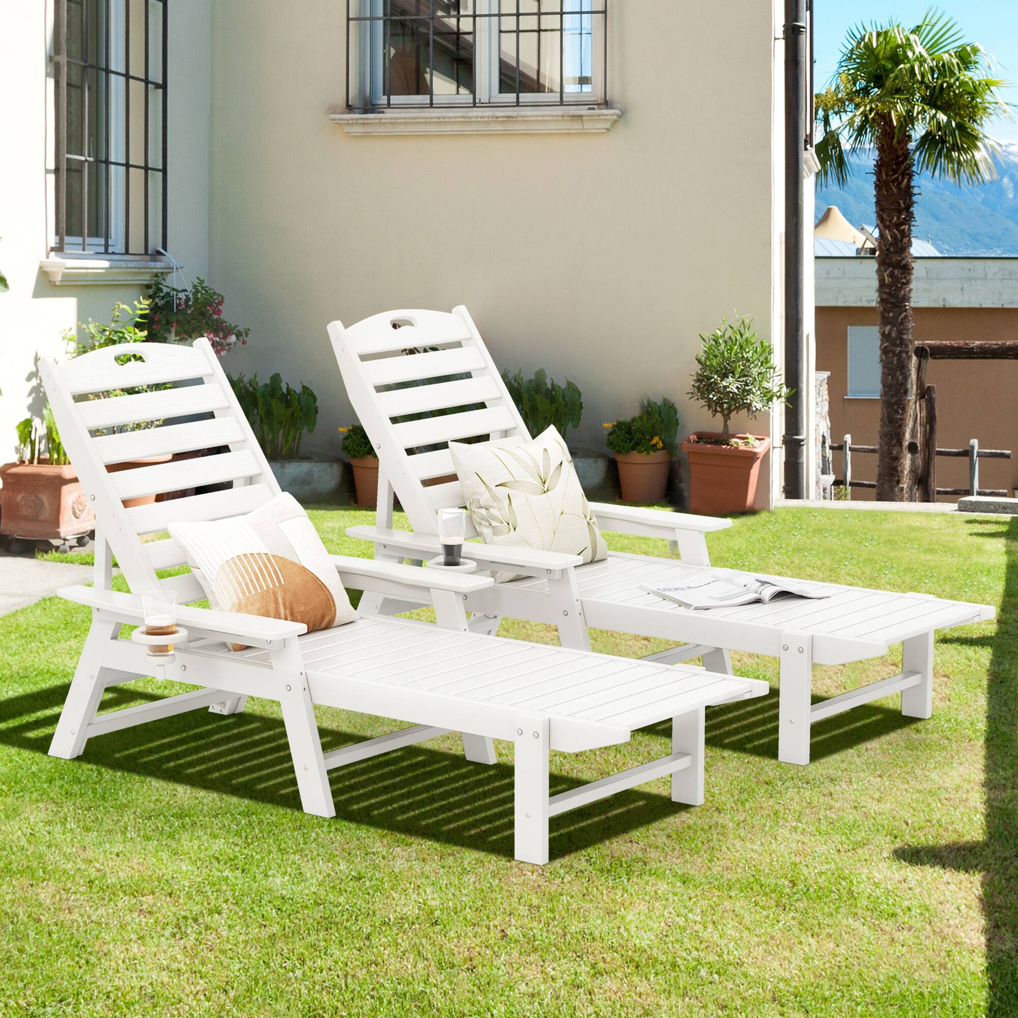 The image does not contain any text. It shows a garden setting with two white wooden sun loungers, a small table, and some potted plants.