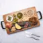 The image features a wooden cutting board with a variety of food items on it. There are several bowls containing different foods, such as pickles, olives, nuts, and cheese. Additionally, there are two knives placed next to the cutting board, likely for slicing and preparing the food. The arrangement of the food items and the presence of the knives suggest that this is a setup for a meal or a snack.