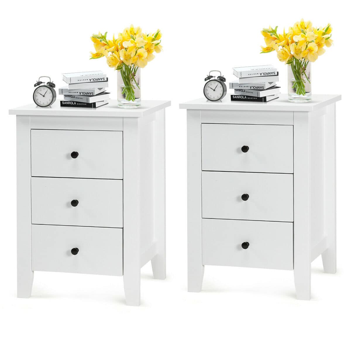 The image features two white dressers with a clock on top of each. The clocks are placed on the left and right sides of the dressers. There are also two vases of flowers, one on each dresser, adding a touch of color and decoration to the scene. Additionally, there are two books placed on the left dresser, possibly serving as reading material or decorative items.