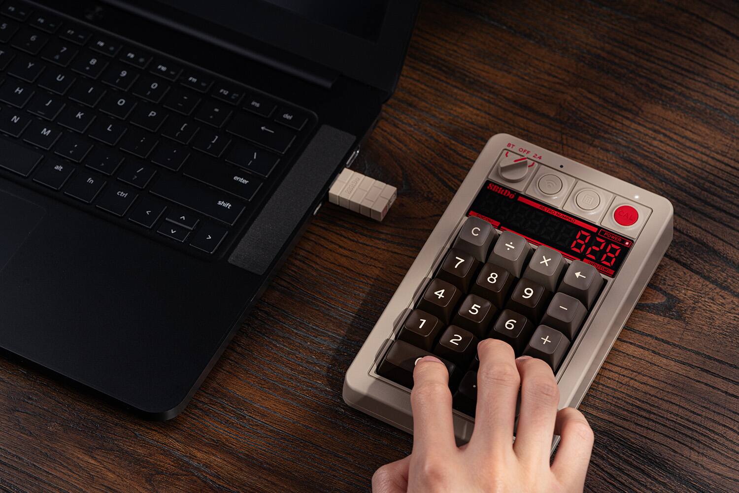 The image shows a person using a calculator with the numbers 1 through 9 displayed on it. The calculator is placed on a wooden table, and the person is typing on the calculator. The calculator is connected to a laptop, which is also on the table. The laptop keyboard is visible, and the person is using the laptop in addition to the calculator.