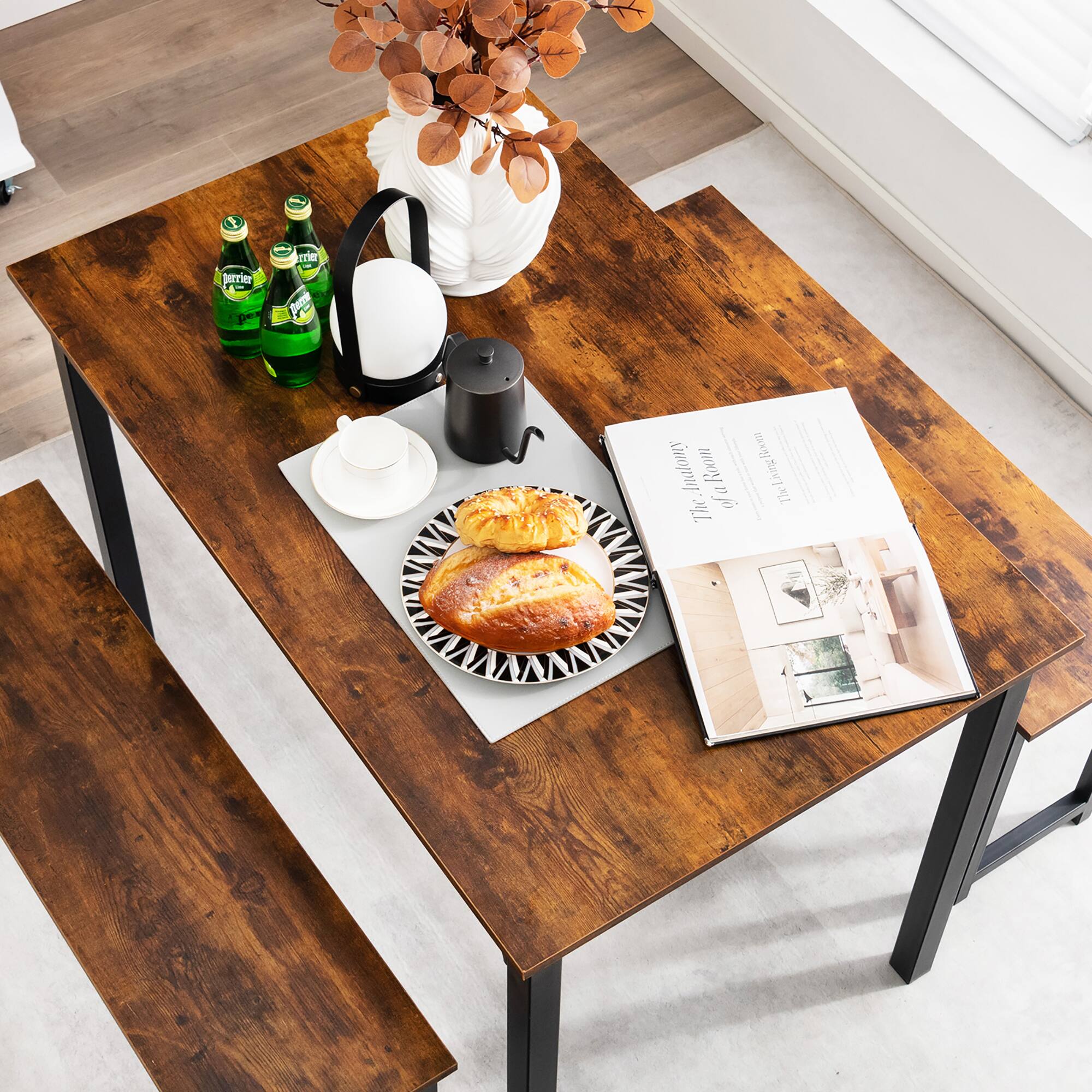 The image shows a wooden table with a plate of bread, a vase of flowers, and a bottle of water. There is also a book on the table, and a cup is placed nearby. The table is situated in a room with a window, providing natural light to the space. The arrangement of the items on the table creates a cozy and inviting atmosphere.