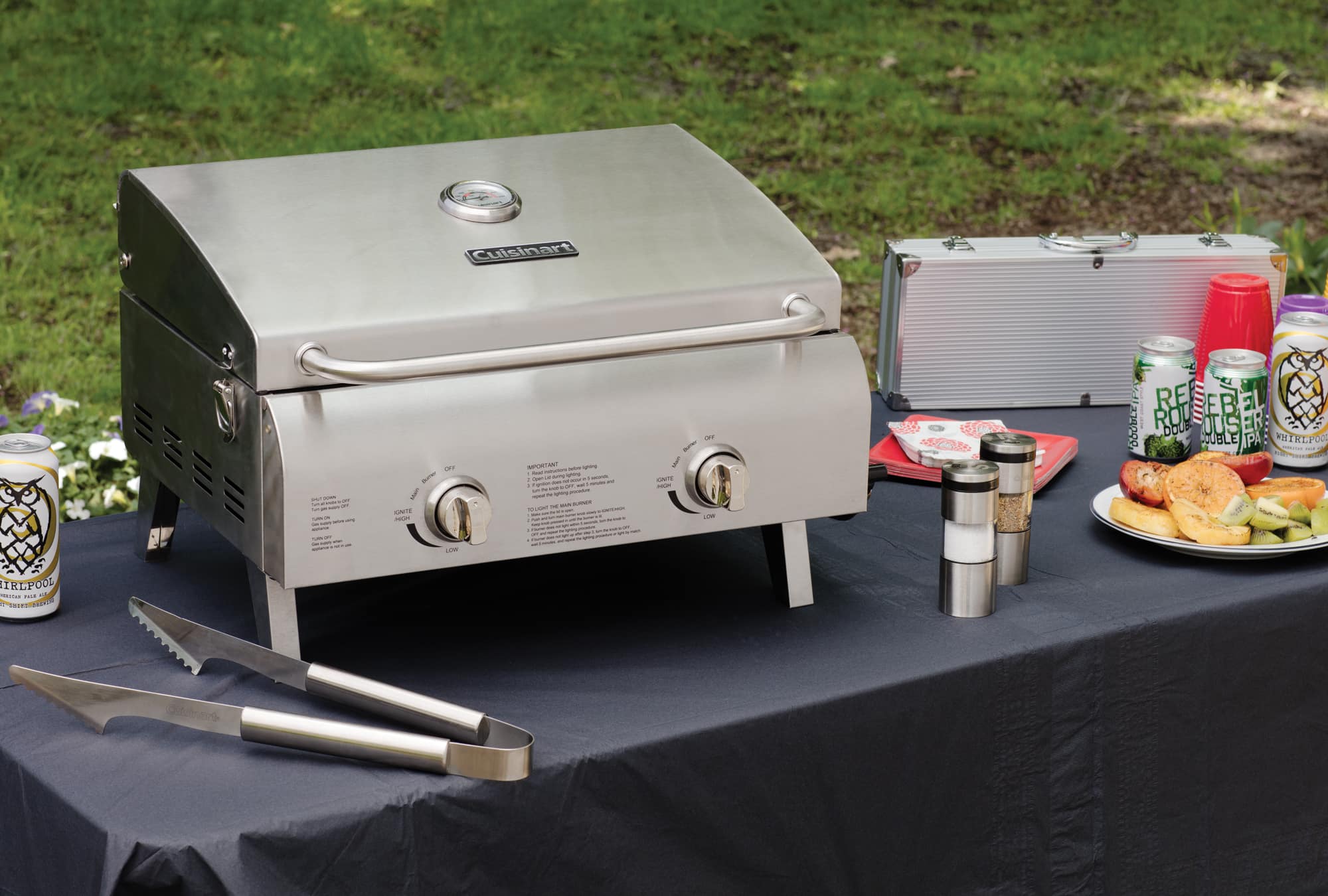 The image shows an outdoor grilling setup with a Weber grill, a table, and various food items and drinks. There is a plate of food, including a sandwich, and a beer on the table. There are also two cups, one near the beer and another further away. A knife is placed on the table, likely for cutting or preparing the food. The scene suggests a casual outdoor gathering or picnic, where people can enjoy grilled food and drinks in a relaxed atmosphere.