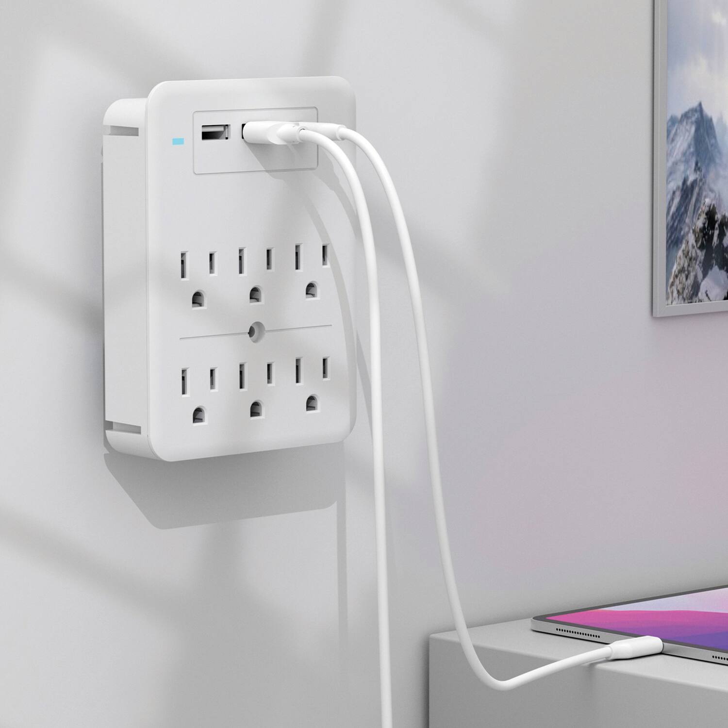 The image shows a white power strip mounted on a wall with multiple outlets and a USB port. A white cable is plugged into the USB port and extends to a device on a table.