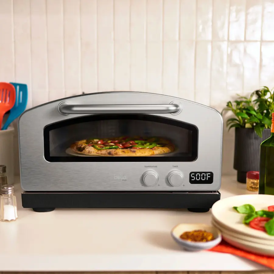 The image shows a silver toaster oven with a pizza inside of it. The oven is placed on a countertop, and there are several items nearby, including a bowl, a bottle, and a spoon. The oven is set to 400 degrees Fahrenheit, and the pizza is cooking inside of it. The scene suggests a casual and cozy atmosphere, with the oven being used to make a delicious meal.