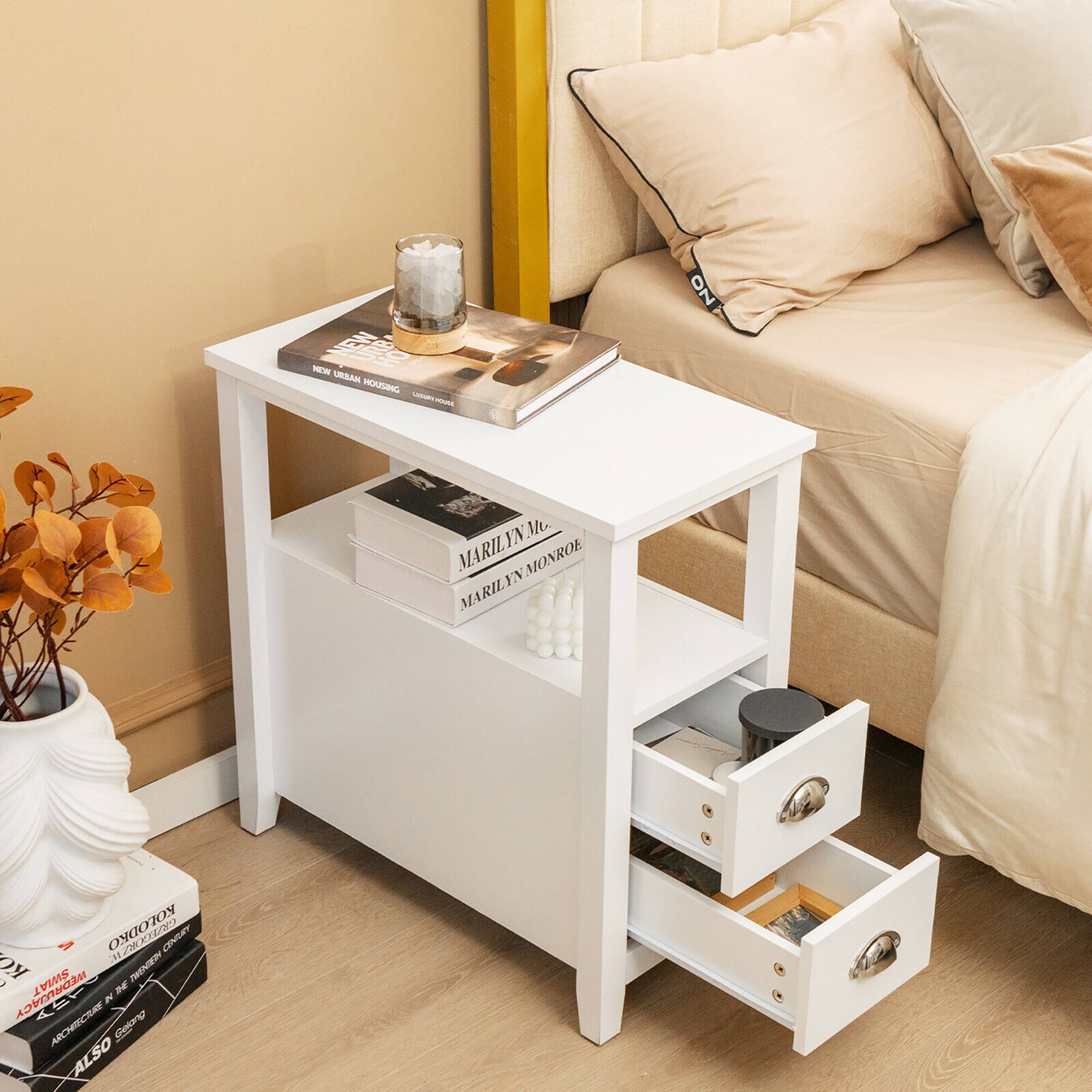 The image features a white table with a candle on it, placed next to a bed. The table has a book on it, and there are two drawers underneath it. Additionally, there is a vase with flowers in it, and a potted plant nearby. The scene appears to be in a bedroom.