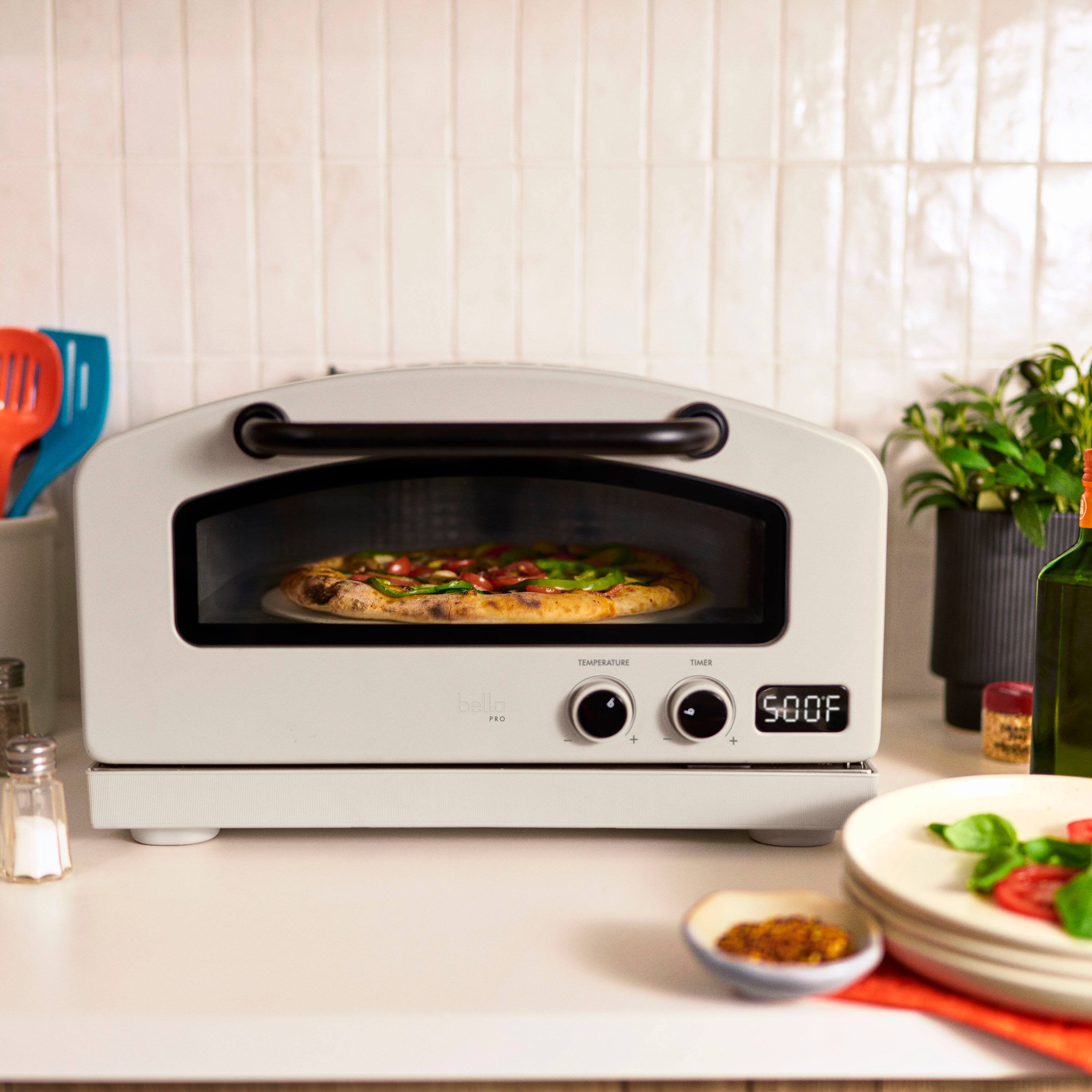 The image shows a pizza inside an oven, which is set to 500 degrees Fahrenheit. The oven is placed on a counter, and there are some potted plants nearby. Additionally, there are two bowls on the counter, one containing vegetables and another with a spoon in it. A bottle is also visible on the counter, possibly containing a beverage or condiment.