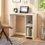 The image shows a small desk with a laptop, a clock, and a bookshelf. The desk is situated in a corner of the room, and there is a chair nearby. The laptop is open, and the clock is placed on the desk. The bookshelf is filled with books, and there is also a vase on the desk. The room appears to be a home office or a workspace, providing a comfortable and organized environment for work or study.