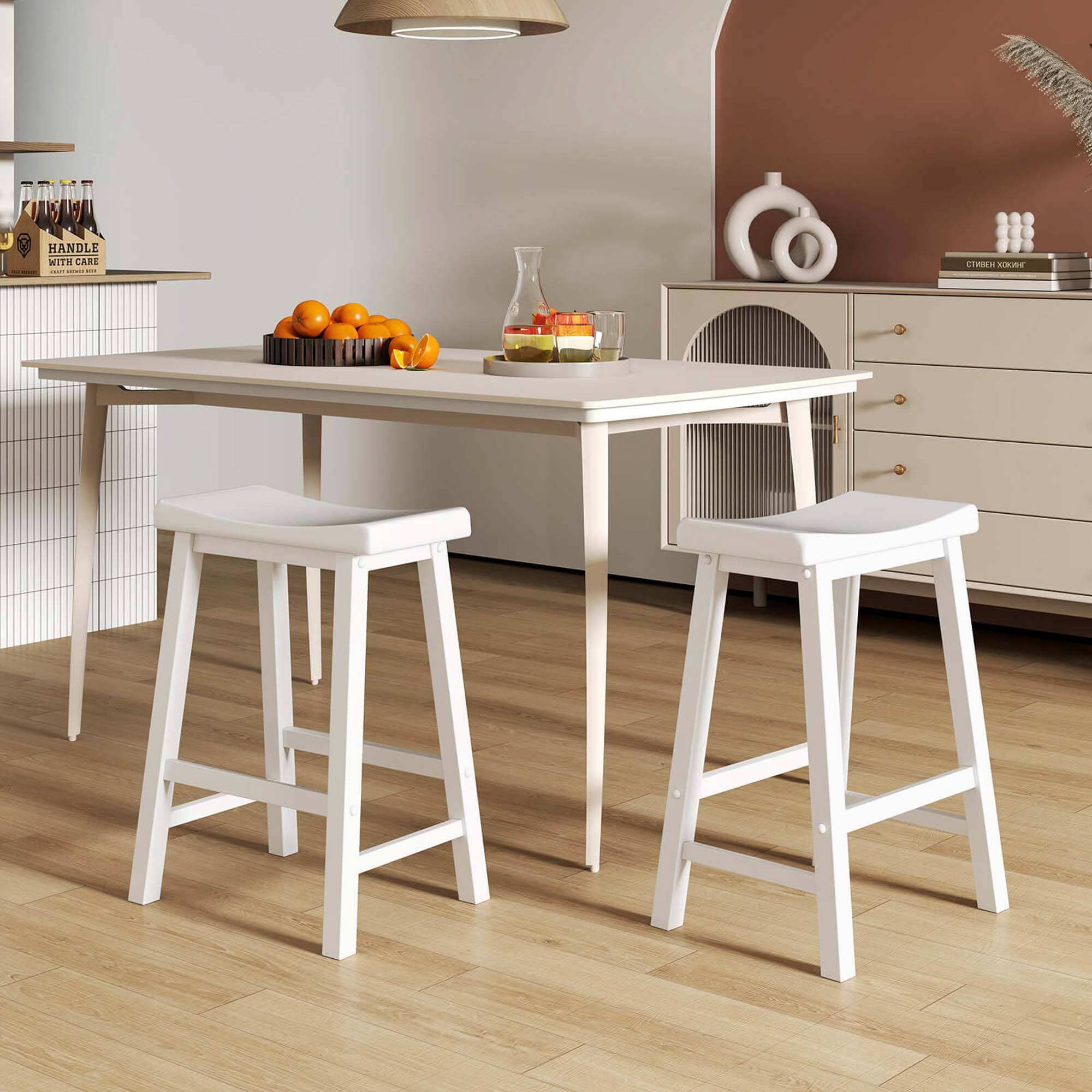 The image features a kitchen scene with a white table and two white stools. The table is set with a bowl of fruit, including apples and oranges. There are also a couple of bottles placed nearby. The kitchen is decorated with a vase and a book, adding to the cozy atmosphere. The overall scene is a pleasant and inviting space for enjoying a meal or spending time with family and friends.