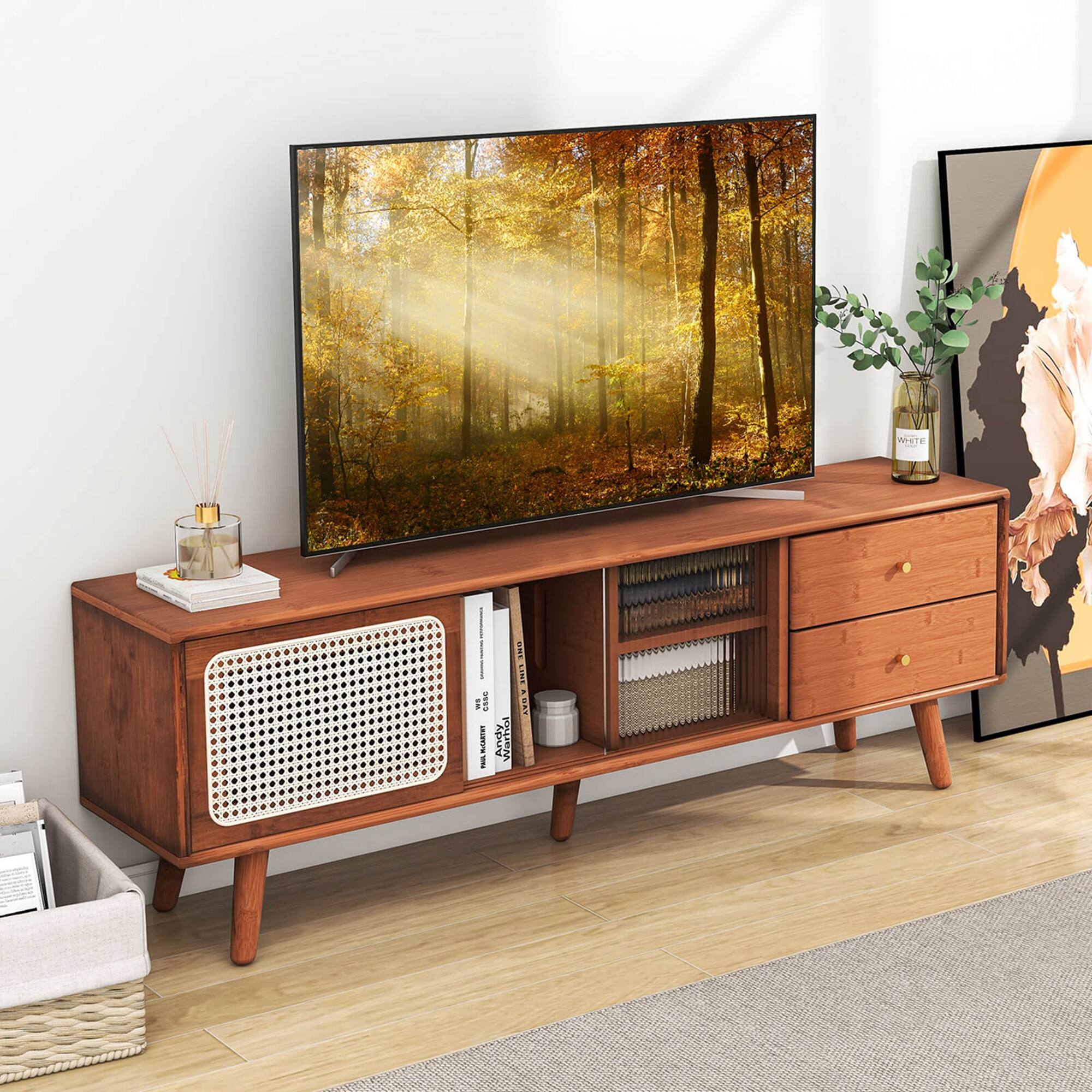 The image features a flat screen TV placed on a wooden stand. The TV is turned on and displaying a picture of trees. The stand also has a drawer and a shelf, and it is situated in front of a wall. There are a few books on the shelf, and a vase with flowers is placed nearby. Additionally, there is a picture hanging on the wall, and a potted plant is located next to the TV.
