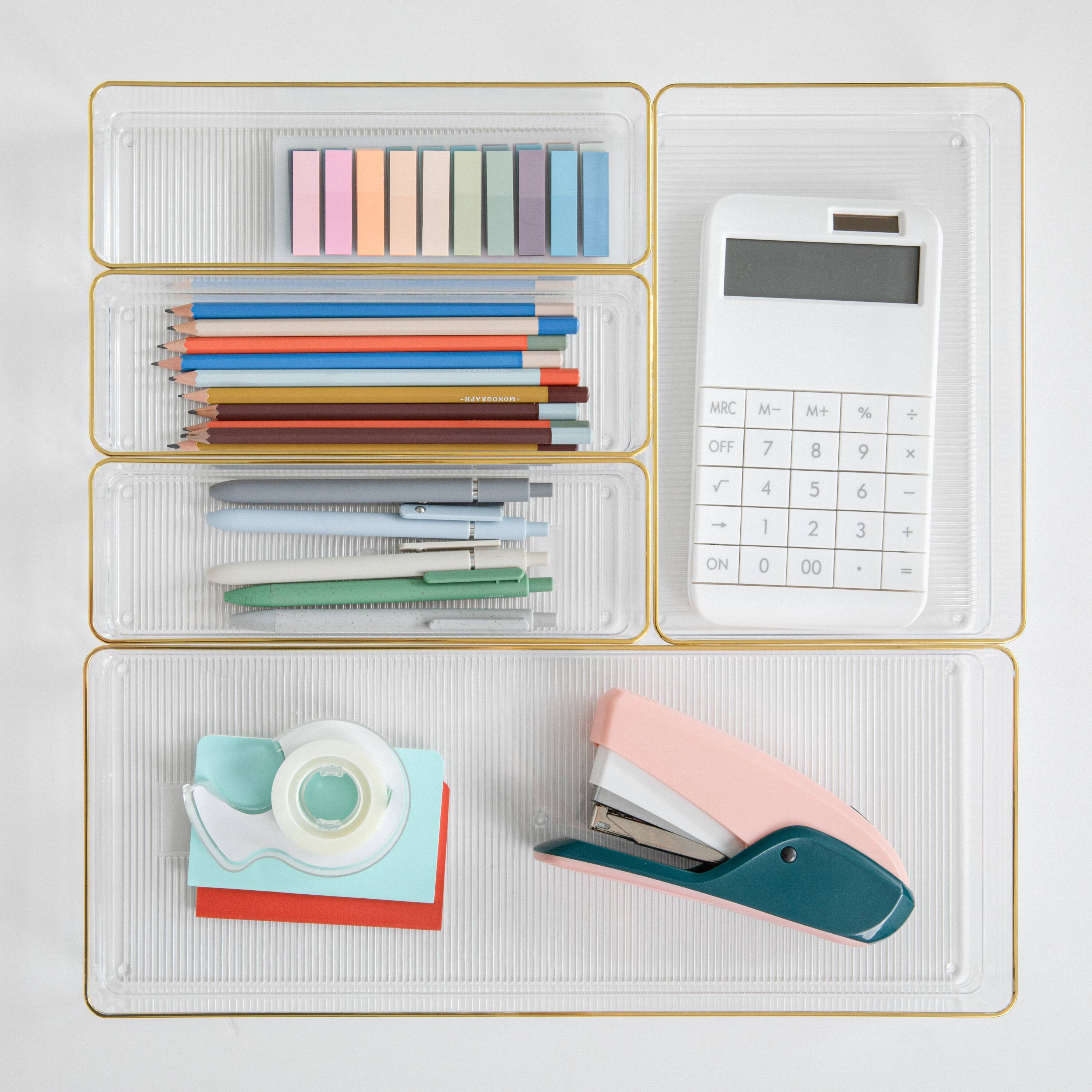 The image shows a clear plastic container with various office supplies, including pens, pencils, and a calculator. The calculator is placed in the top right corner of the container, while the pens and pencils are arranged in different sections. The container is filled with these items, making it a convenient and organized storage solution for office supplies.