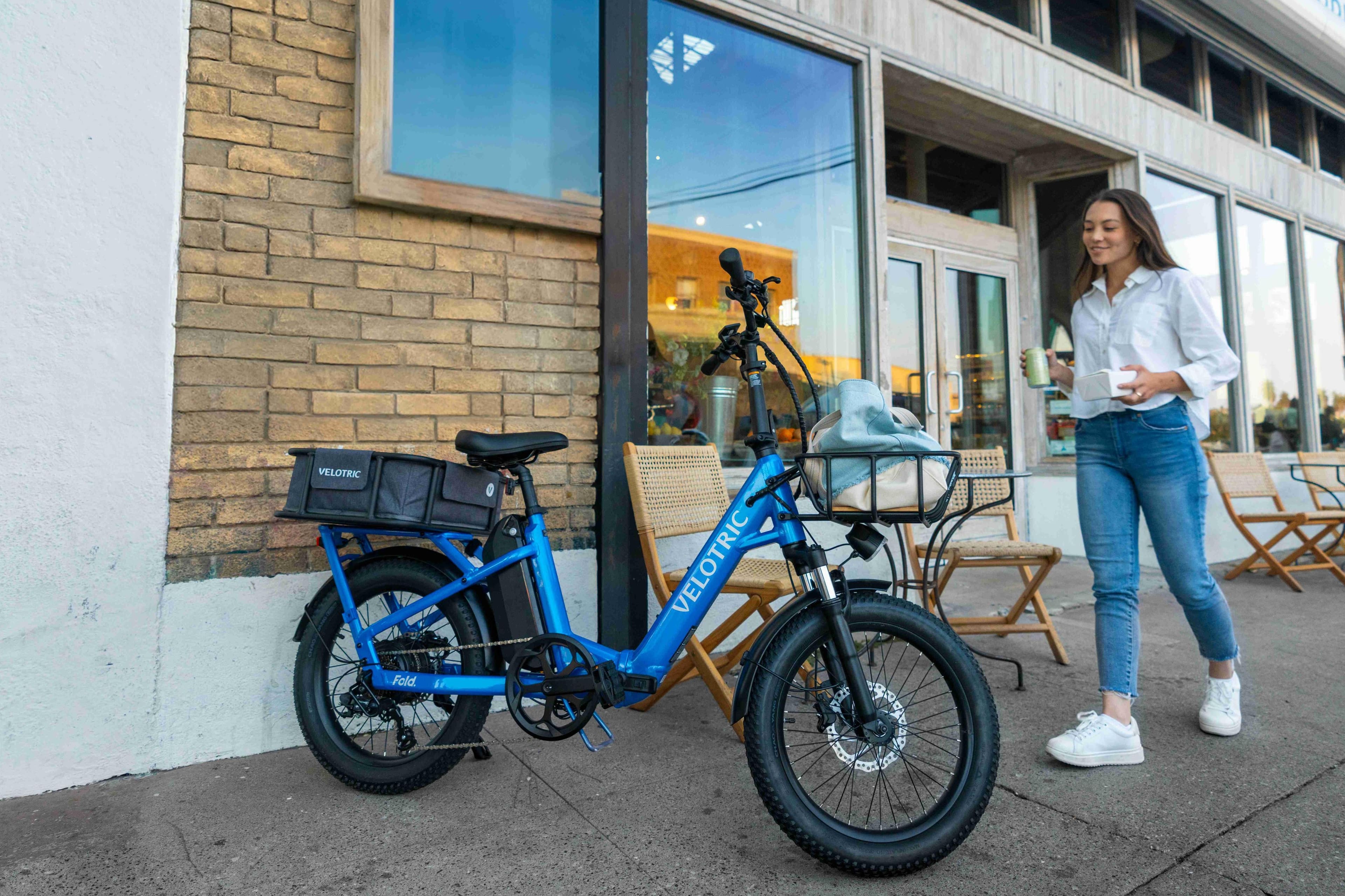 The image shows a woman standing next to a blue foldable bicycle, which is parked outside a building. The bicycle is a Velotric foldable bike, and the woman appears to be preparing to ride it. The scene also includes a few chairs and a dining table nearby, suggesting that the location might be a cafe or a similar outdoor seating area.