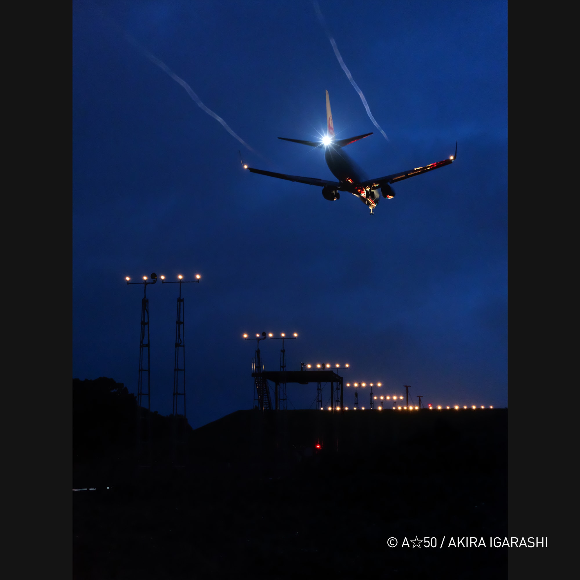 Akira Igarashi took this photo of a plane flying over a runway at night. The image captures the airplane in flight, with its lights on, as it soars through the sky. The scene is set against a backdrop of a dark night, creating a dramatic and captivating image.