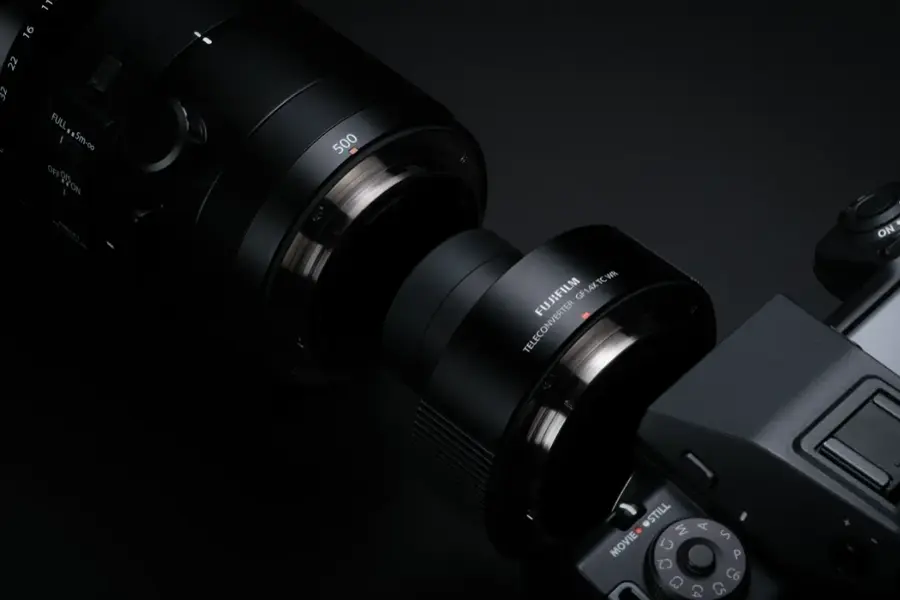The image features a close-up of a camera lens, specifically a Fujifilm GF14XTCWR teleconverter lens. The lens is attached to a camera, and it is positioned next to another lens. The focus of the image is on the lenses, showcasing their details and features.