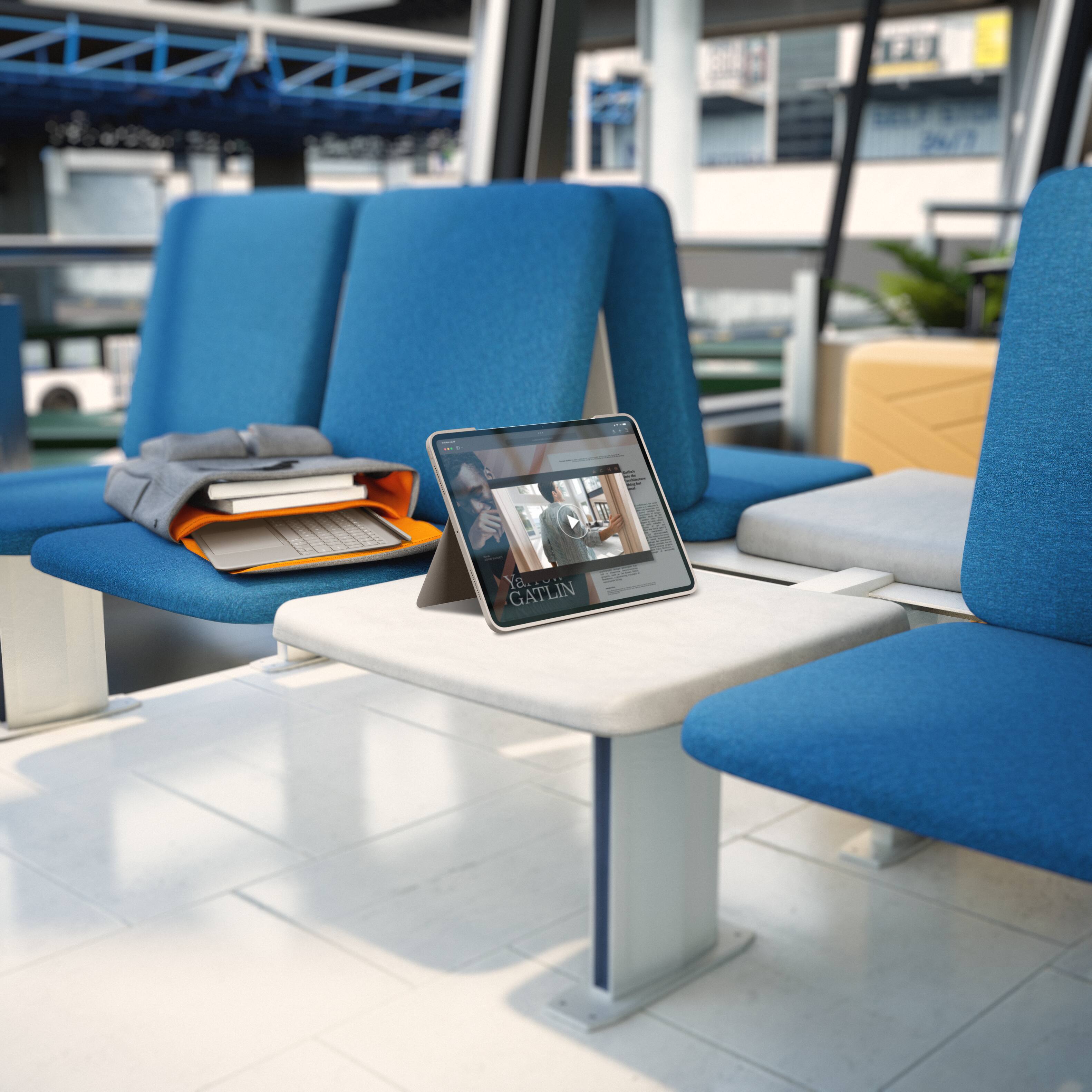 The image shows a table with a laptop and a book on it. The table is surrounded by blue chairs, and there is a potted plant nearby. The laptop is open and displaying a video, and the book is placed next to it. The scene appears to be in a waiting area or a public space where people can use the laptop and read the book while sitting on the chairs.